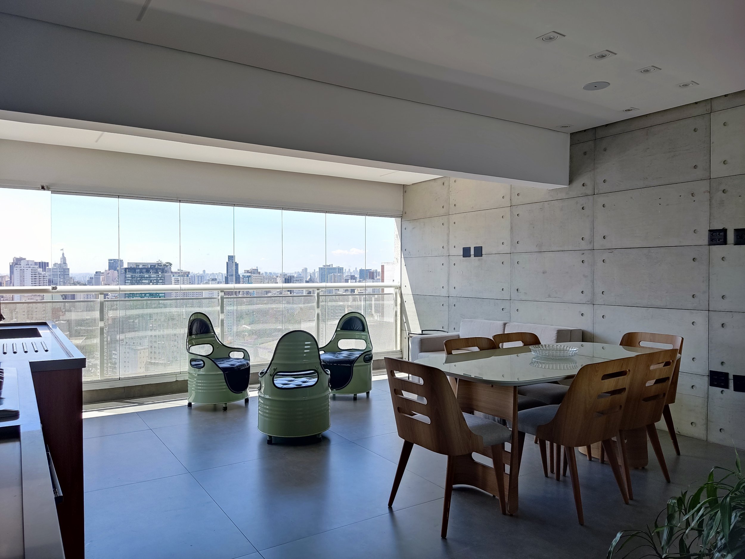 Sala de estar moderna com mesa de jantar de madeira, cadeiras acolchoadas e cadeiras modernas ao lado de uma parede de concreto, com grande janela de vidro oferecendo vista da cidade.