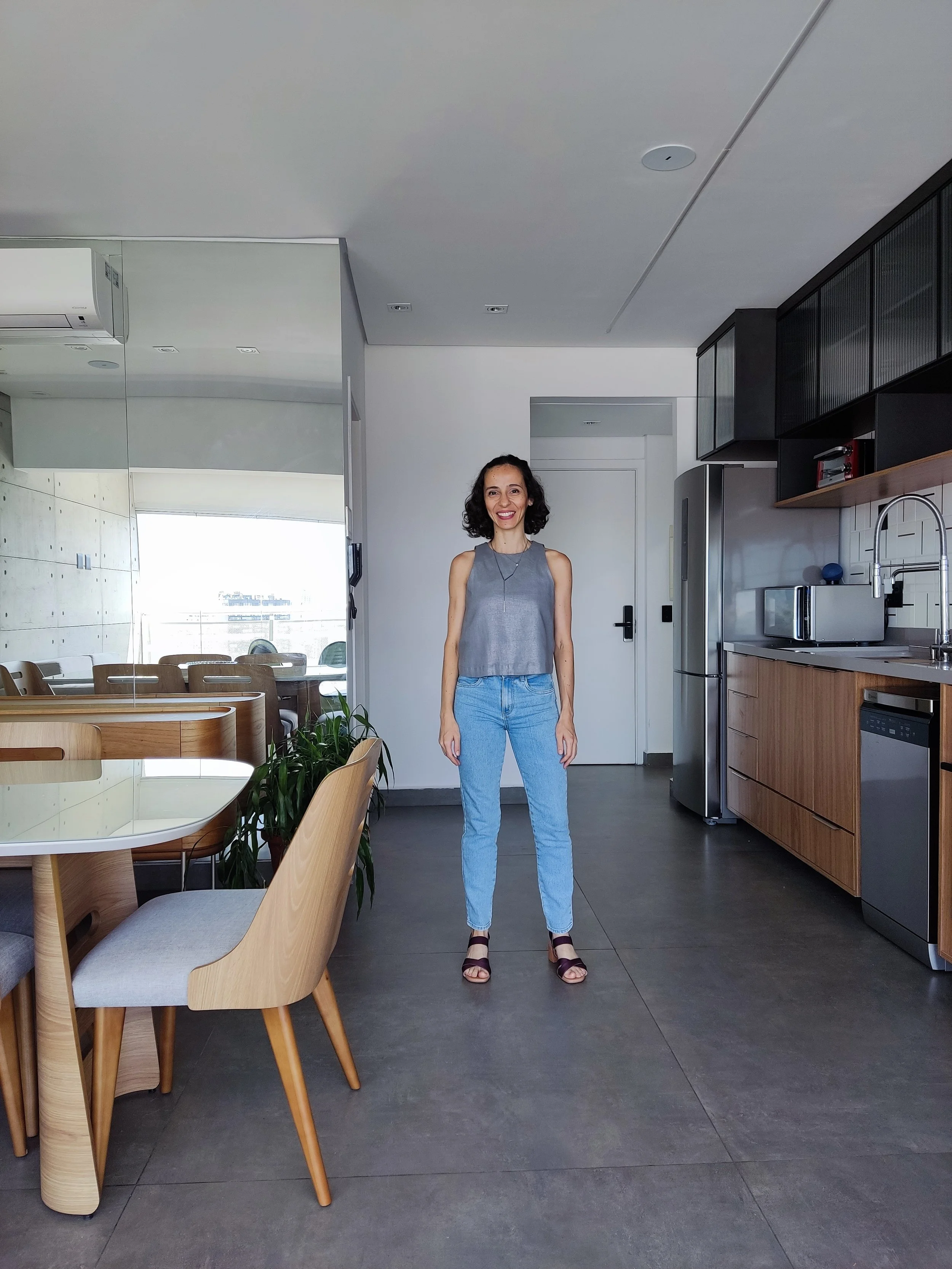 Mulher sorridente de cabelo cacheado escuro, vestindo blusa cinza, jeans claros e sandálias, posando em uma cozinha moderna com móveis de madeira e eletrodomésticos de aço inox.