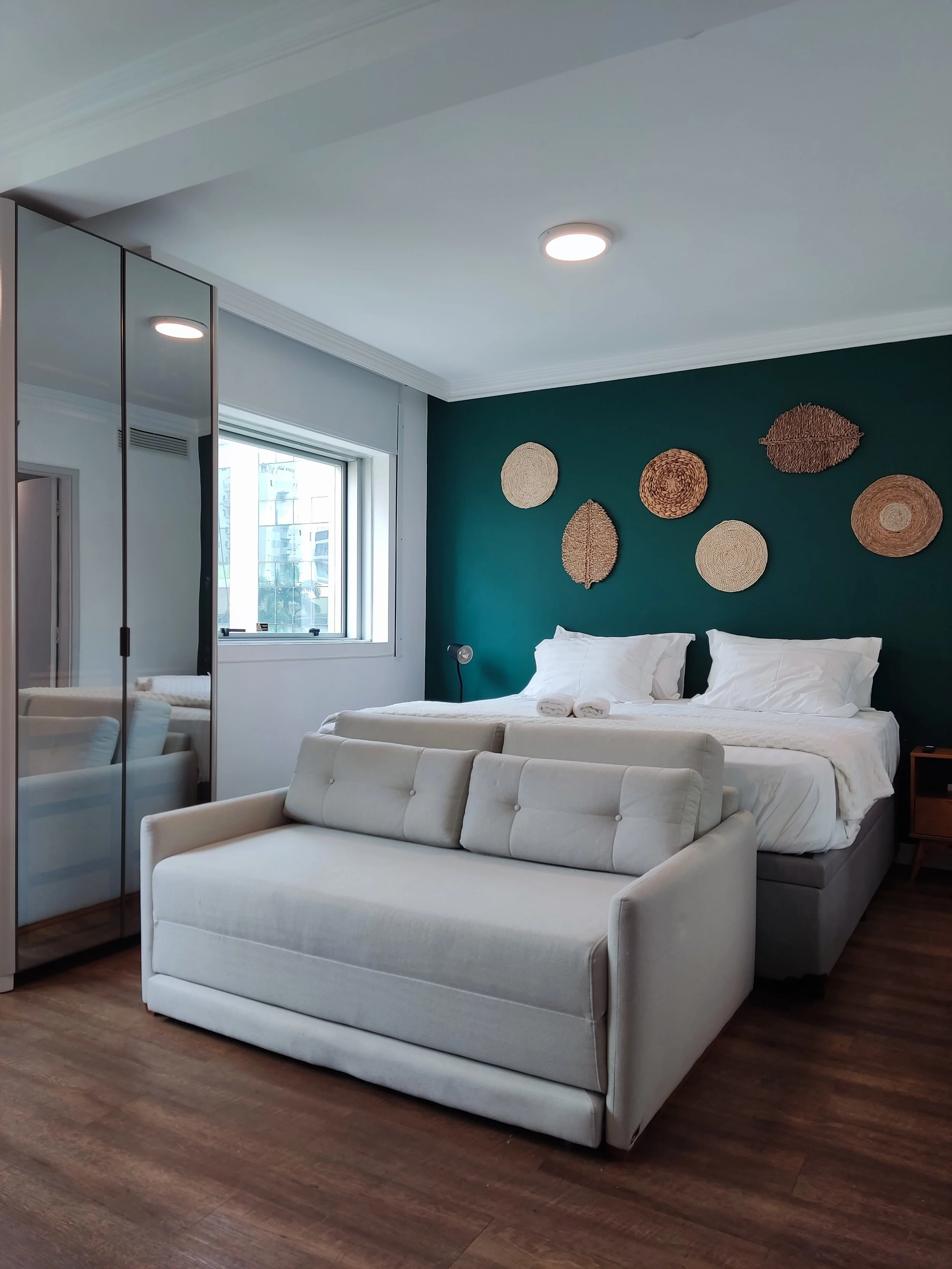 Quarto de hotel com cama de casal, sofá cinza, parede verde com decorativas de palha, janela com vista para prédios, armário espelhado, luminária de leitura ao lado da cama, piso de madeira.