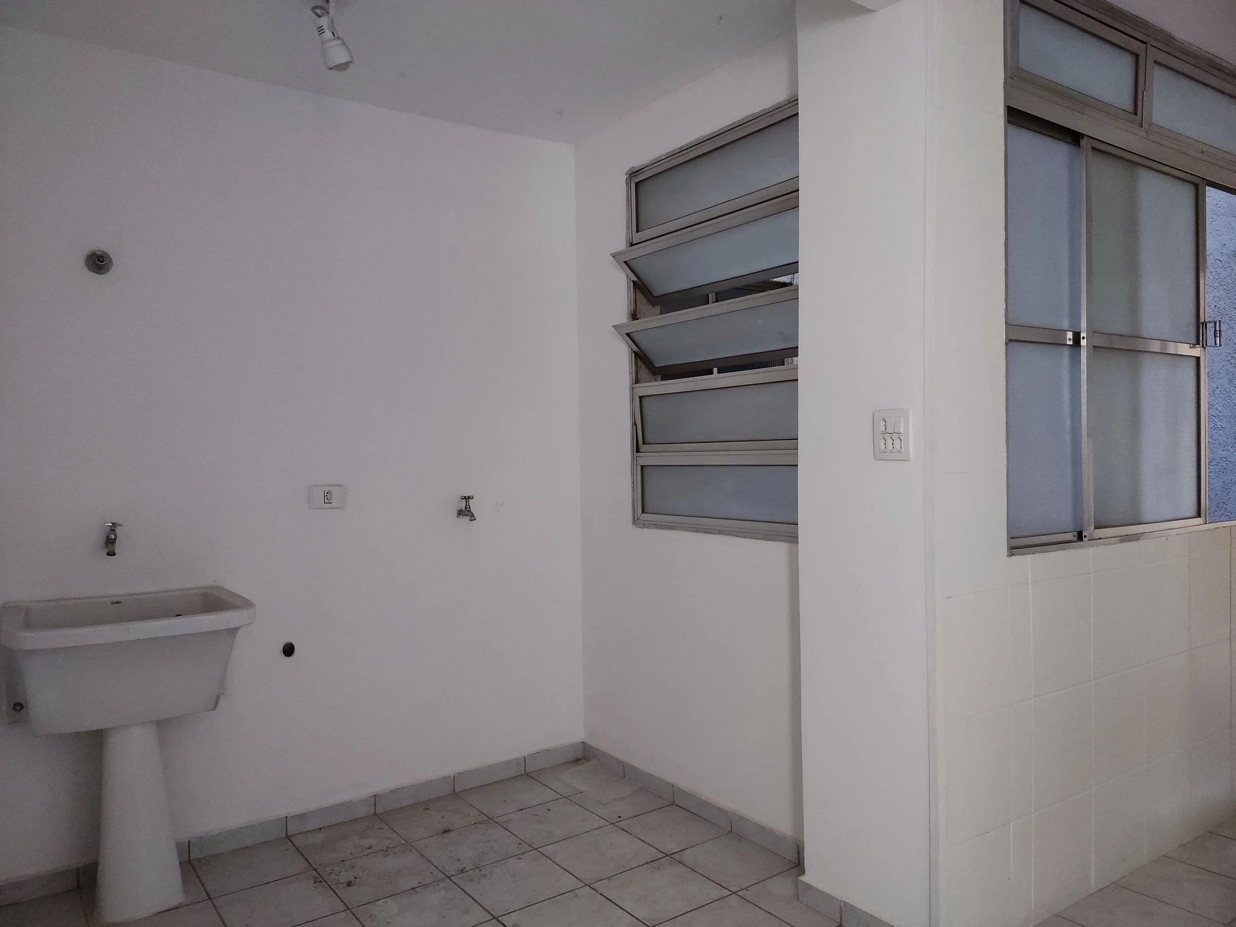 Lavabo branco com torneira, parede branca, piso de azulejo, janelas de vidro com venezianas e interruptor de luz.