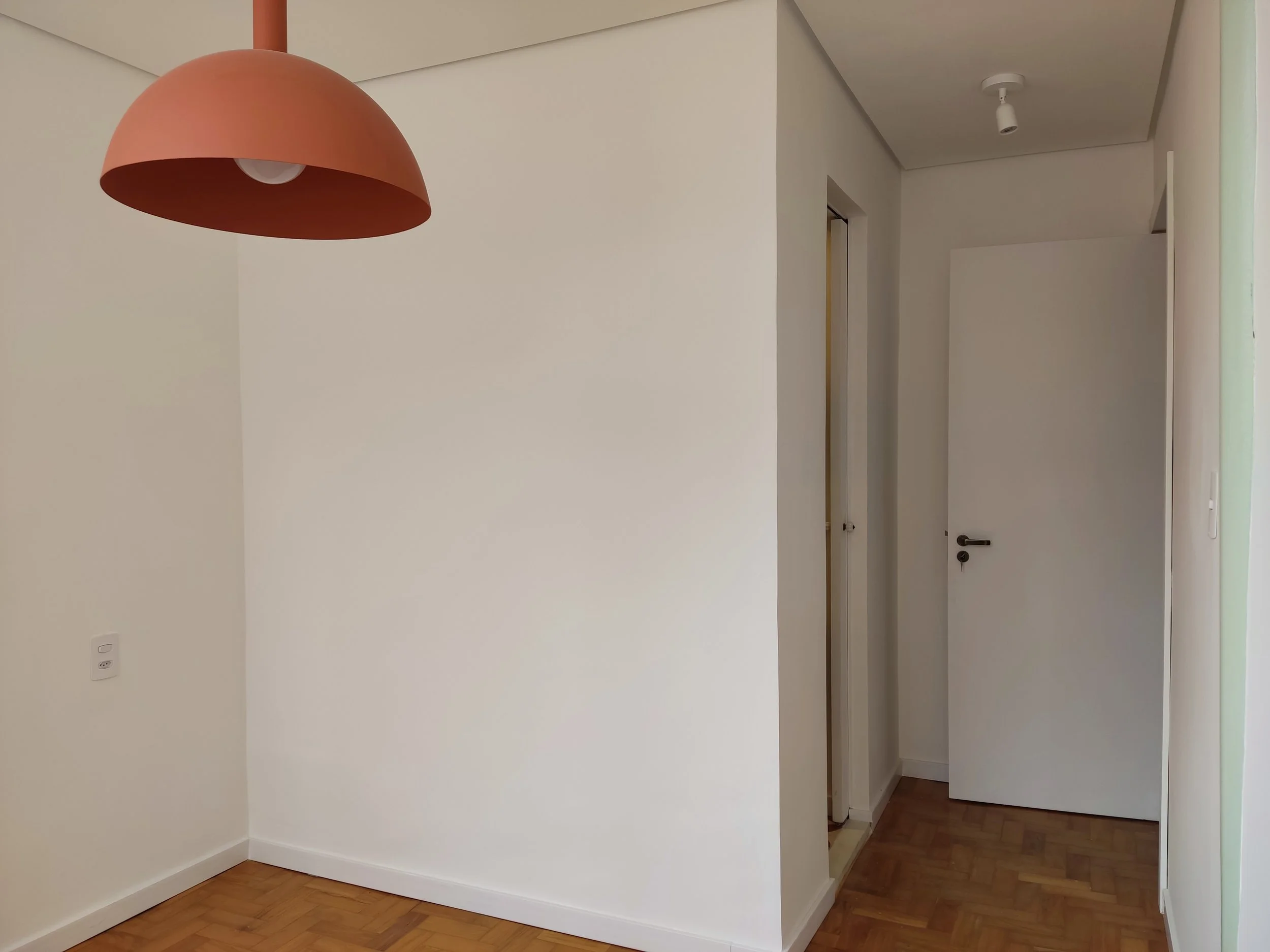 Sala com parede branca, piso de madeira, luminária de tom terracota, porta branca aberta e porta fechada ao fundo.