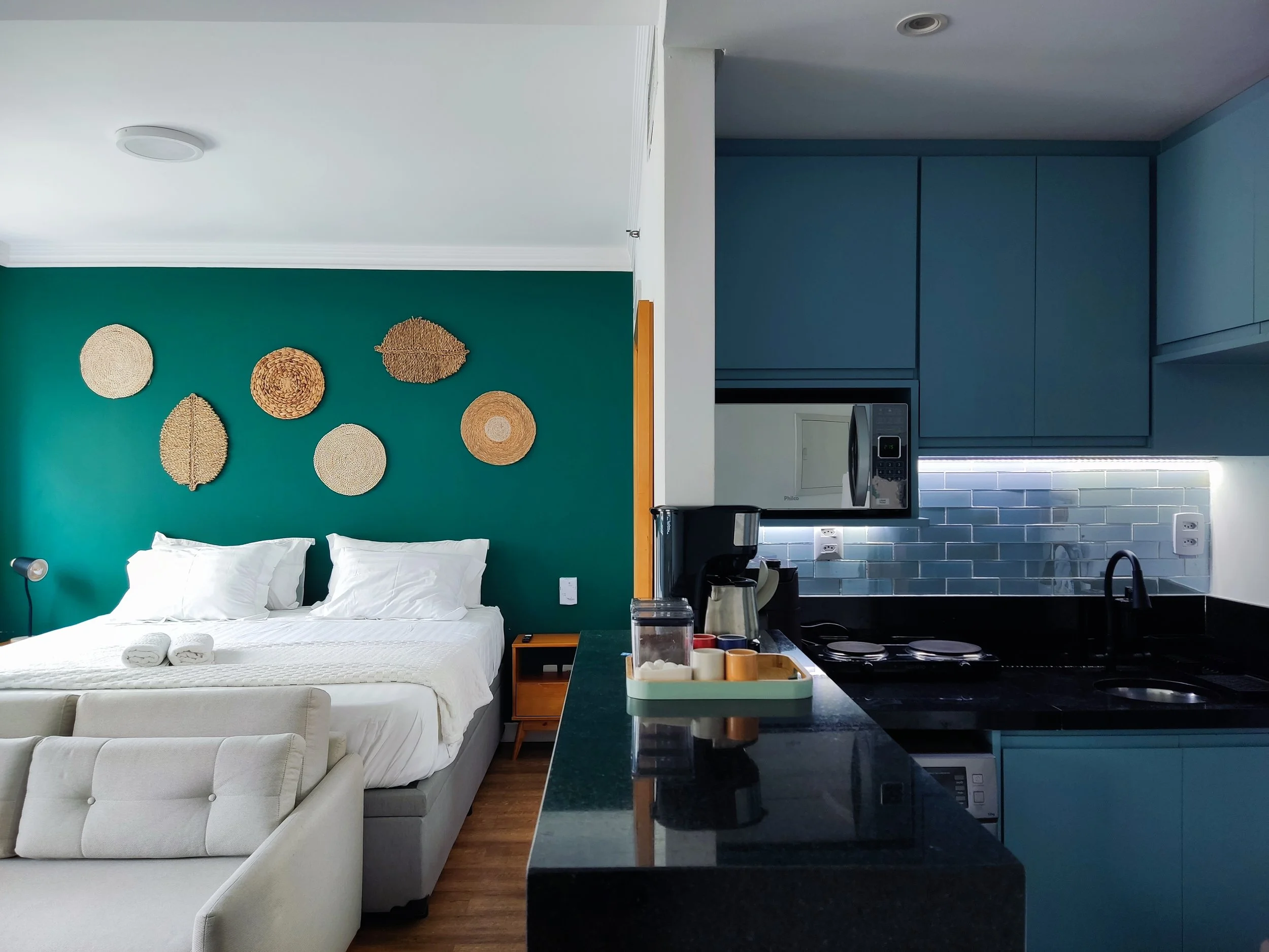 Quarto de hotel com cama de casal, parede verde com decoração de cestos de palha, ao lado um sofá bege, na cozinha azul com backsplash de azulejos cinza, misturador preto e eletrodomésticos.