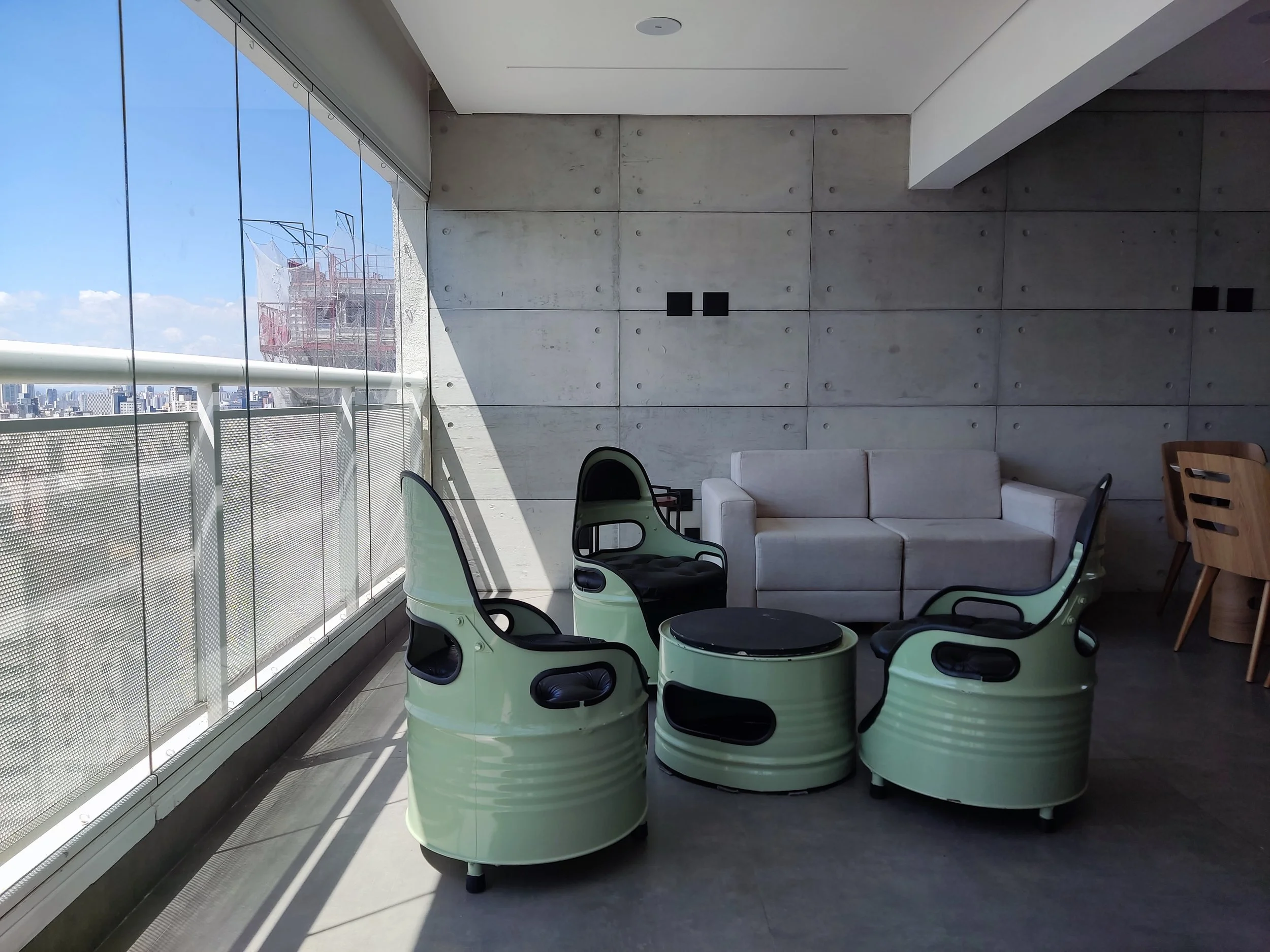 Sala de estar com sofá branco, duas poltronas verdes com detalhes pretos, mesa redonda preta, e uma bancada de jantar de madeira ao lado. Janelas amplas deixam entrar luz natural e mostram um horizonte urbano com edifícios.