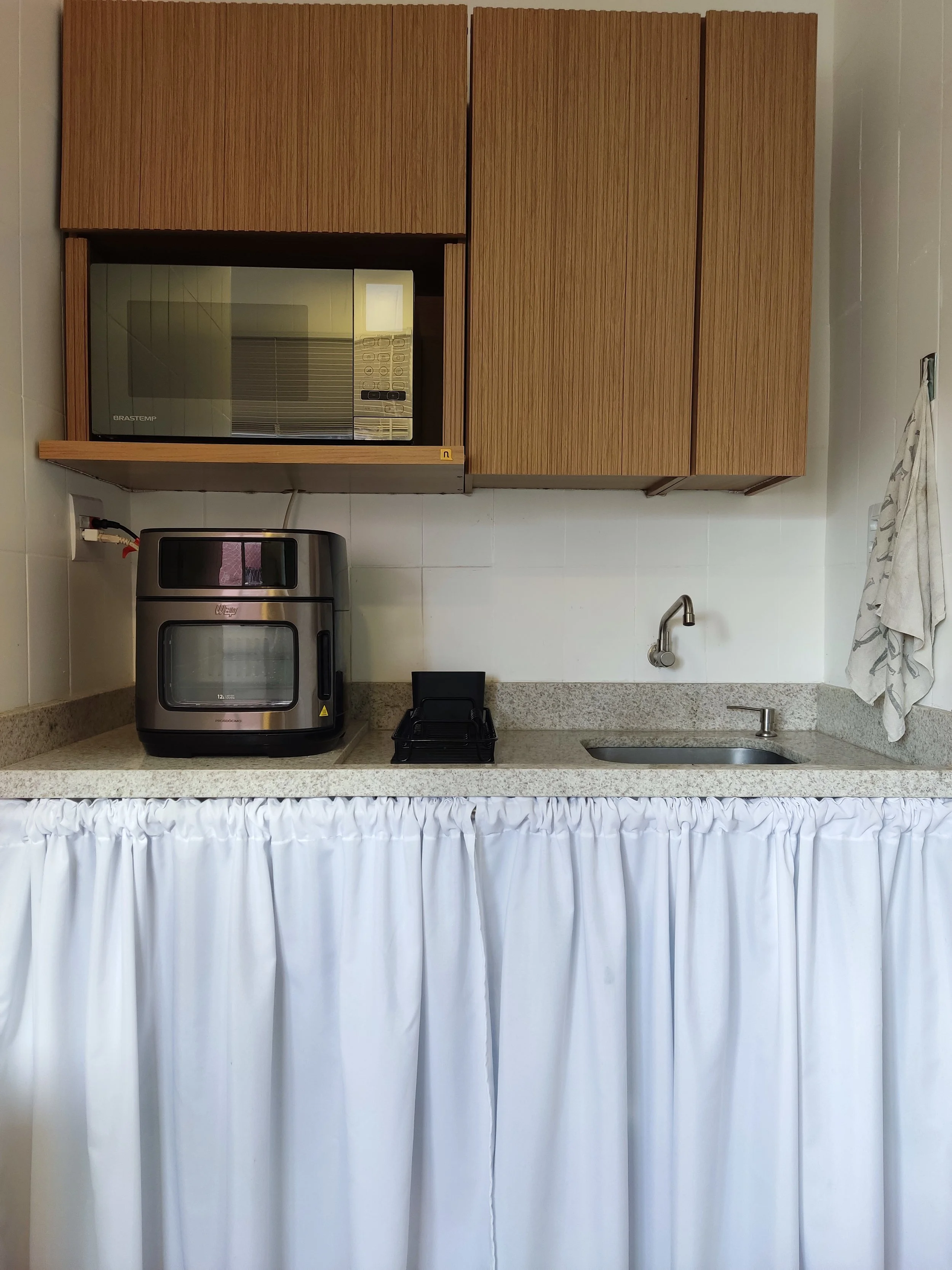 Cozinha com armários de madeira, micro-ondas, forno elétrico, pia, torneira, toalha de tecido branco na frente do balcão.