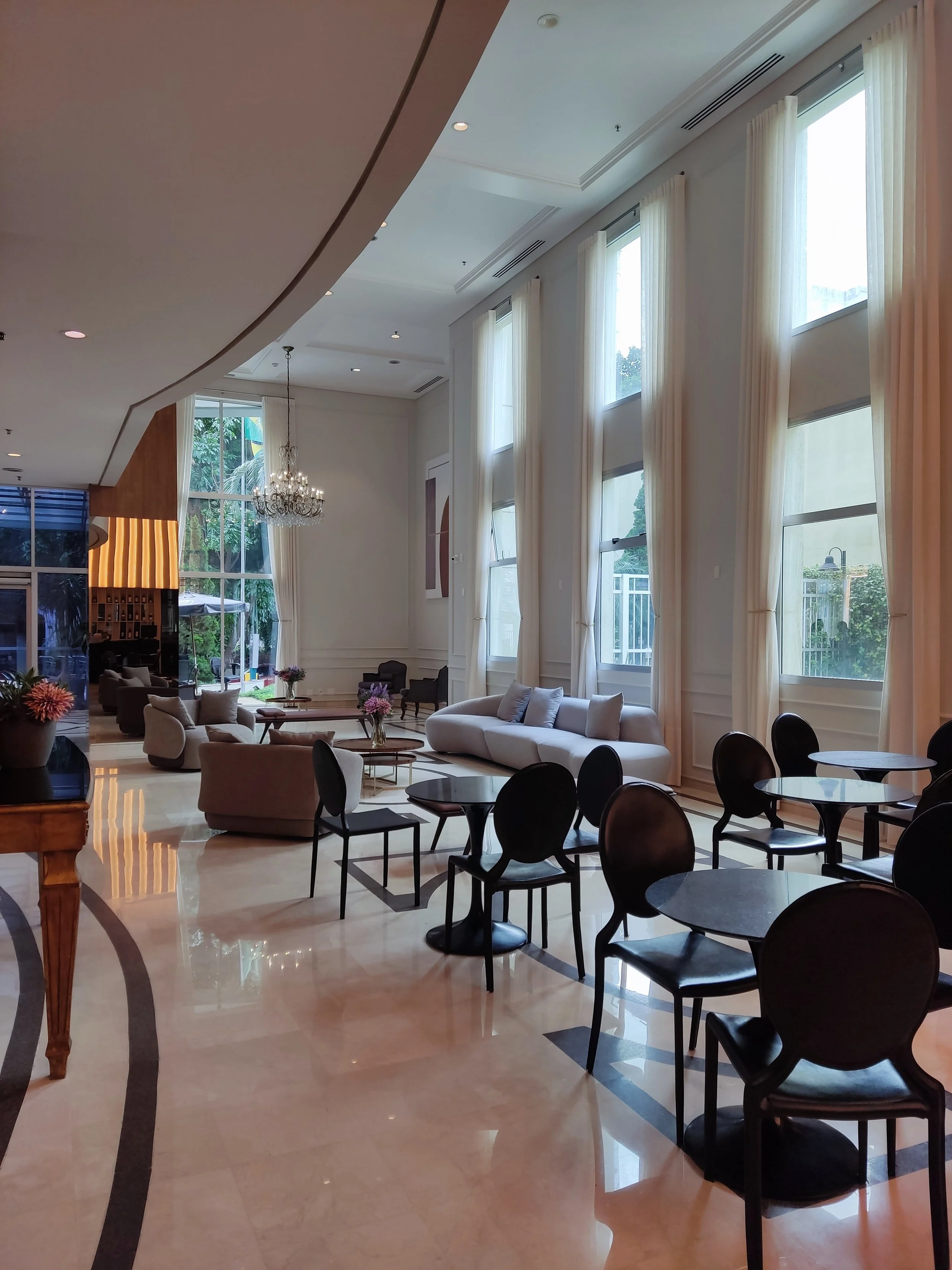 Salão de estar com móveis confortáveis, grandes janelas com cortinas brancas, lustre de cristal e piso de mármore.