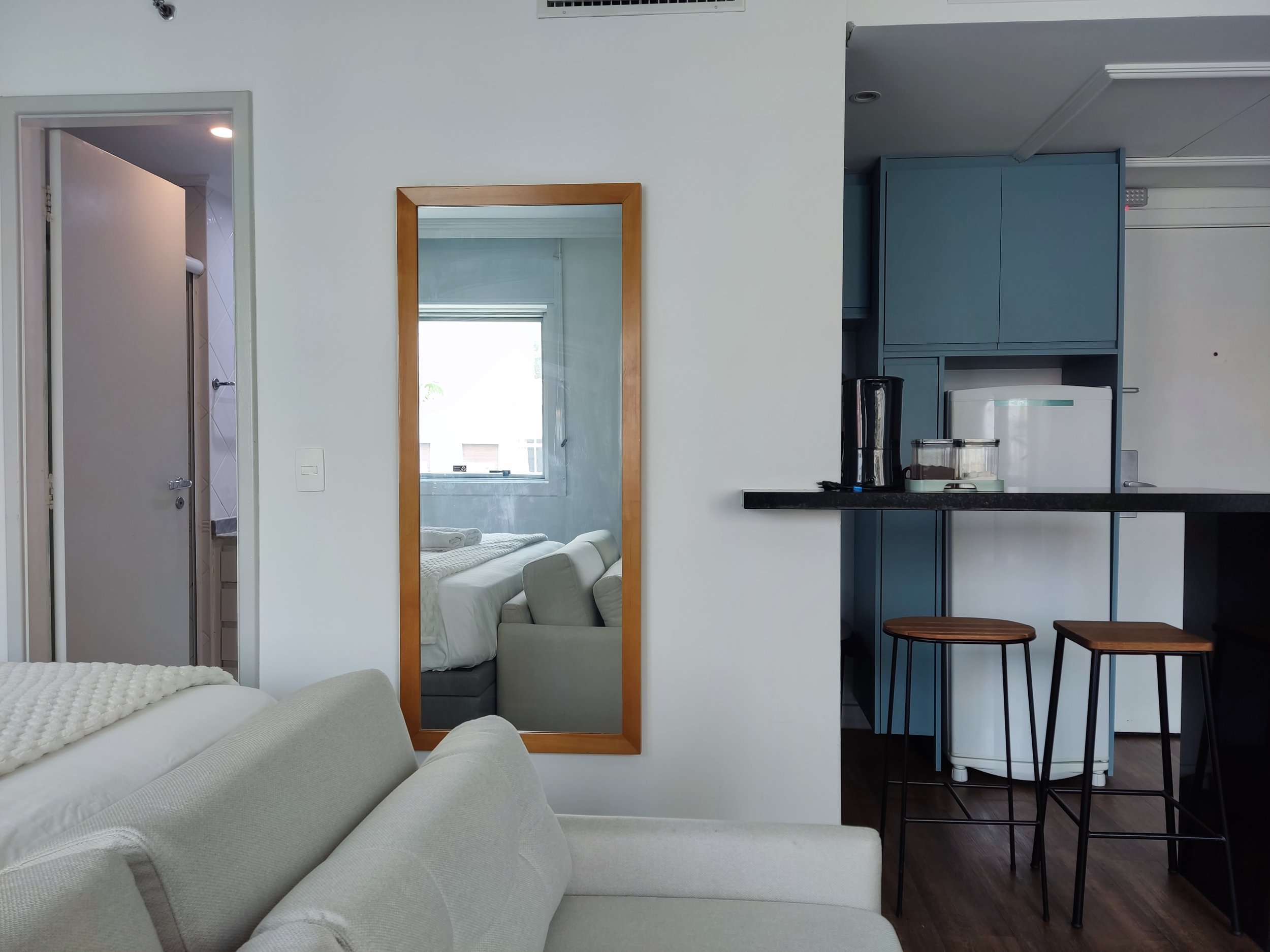Sala de estar com móveis brancos, espelho de madeira na parede, cozinha com armários azuis e bancada preta, duas banquetas de madeira e barista na cozinha. Reflexo do sofá na parede