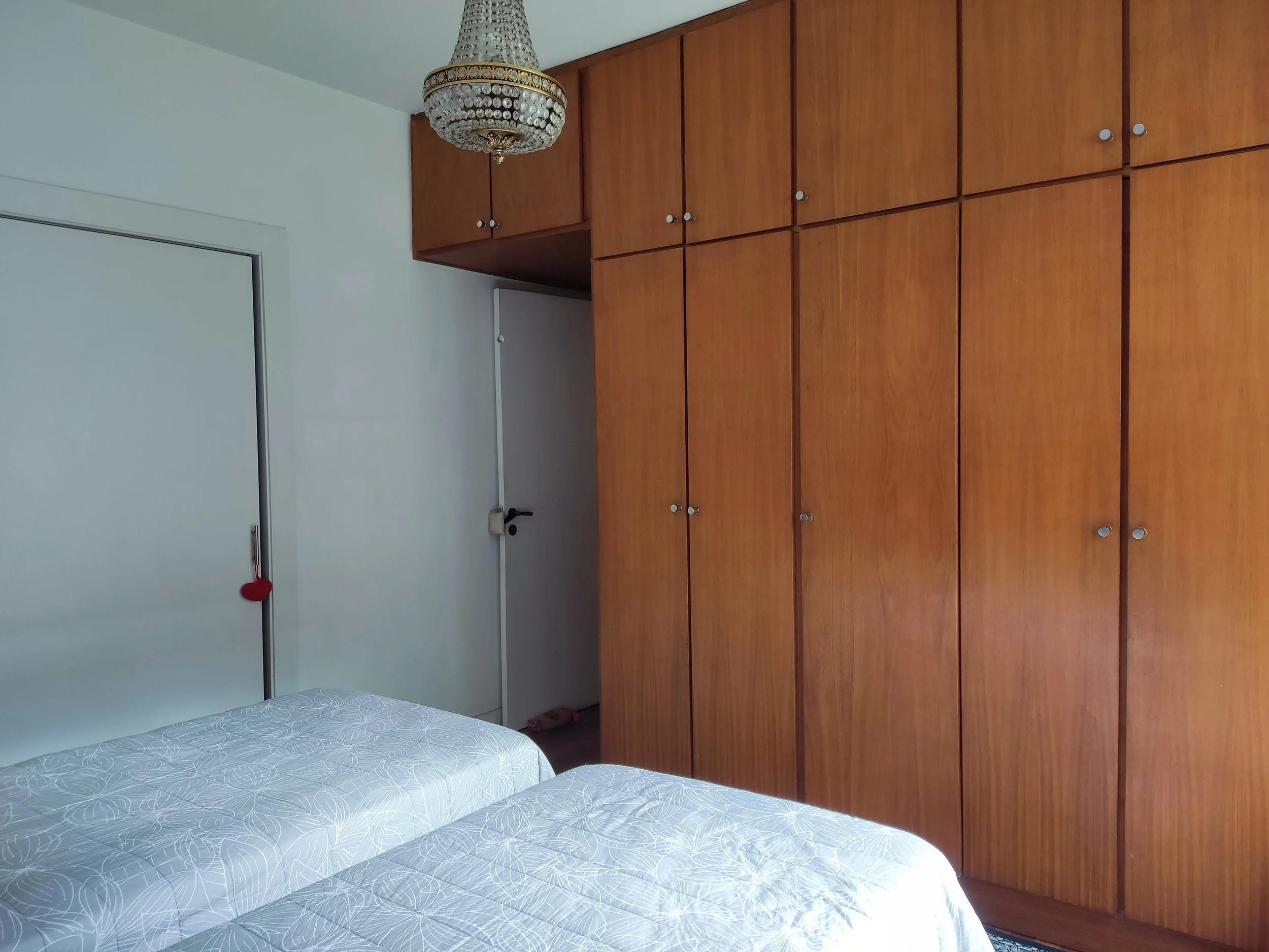 Quarto com armário de madeira ao lado da cama, duas camas de casal com roupas de cama claras, porta branca com coração vermelho pendurado e um candelabro de cristal no teto.