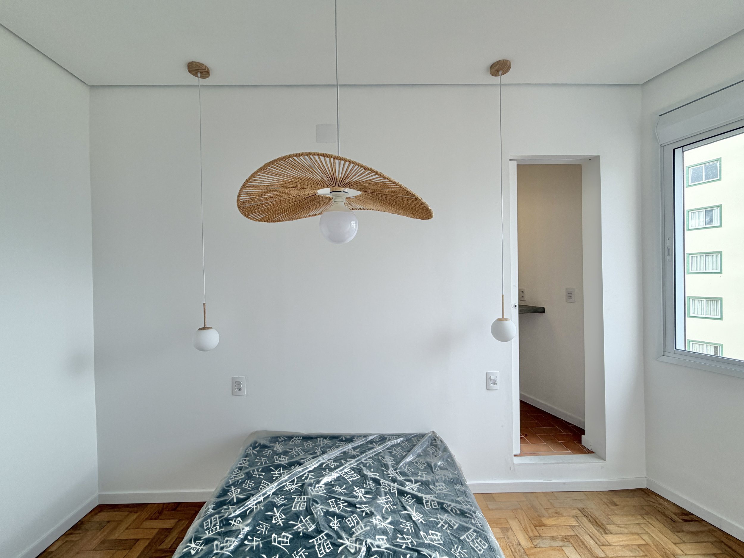 Quarto vazio com piso de madeira, cama com colchão ainda embrulhado, duas luminárias penduradas no teto com acabamento de palha, janela grande com vista para outros prédios e porta para área de serviço.