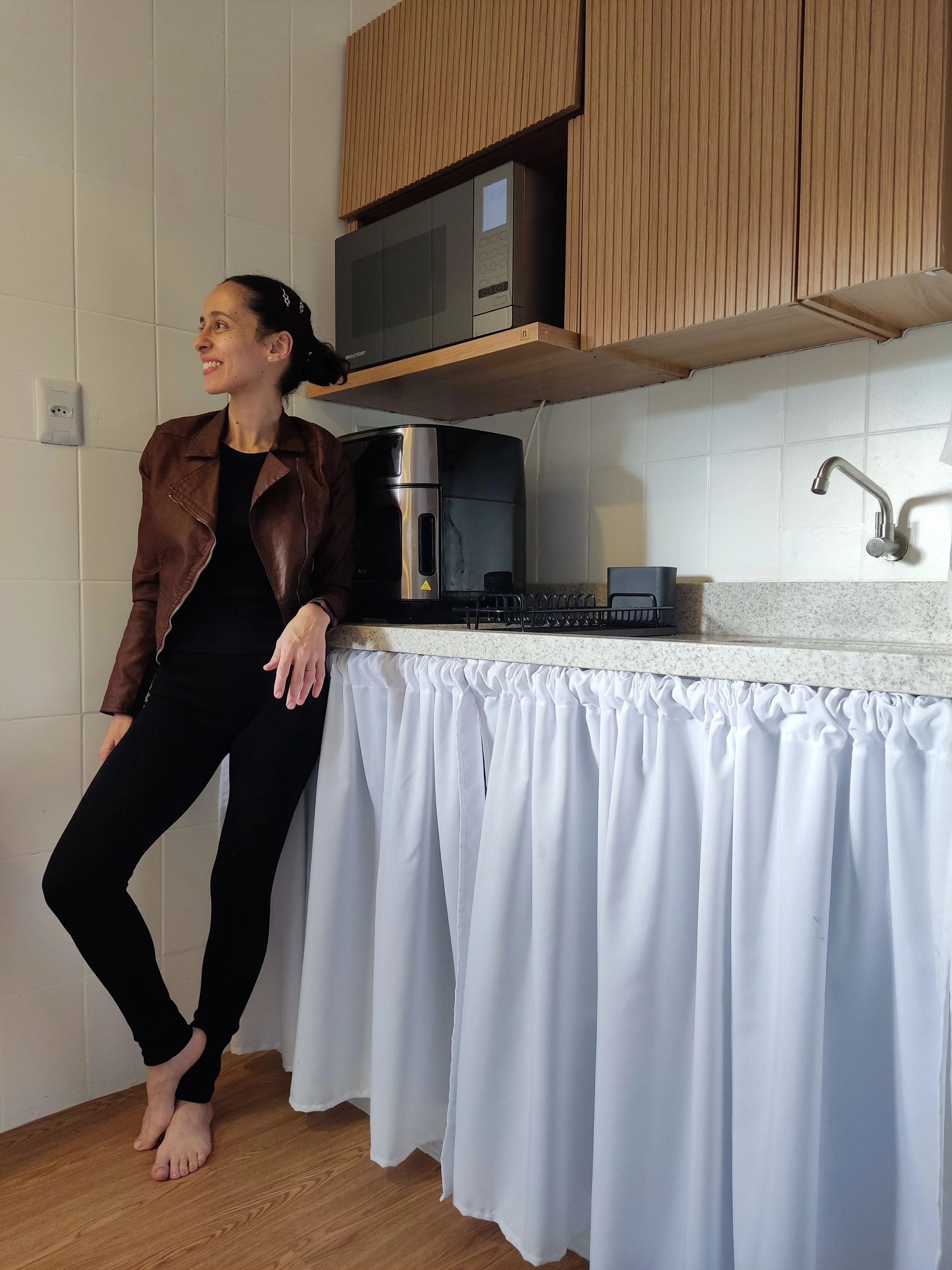 Mulher sorrindo ao lado de bancada de cozinha com micro-ondas e cafeteira.