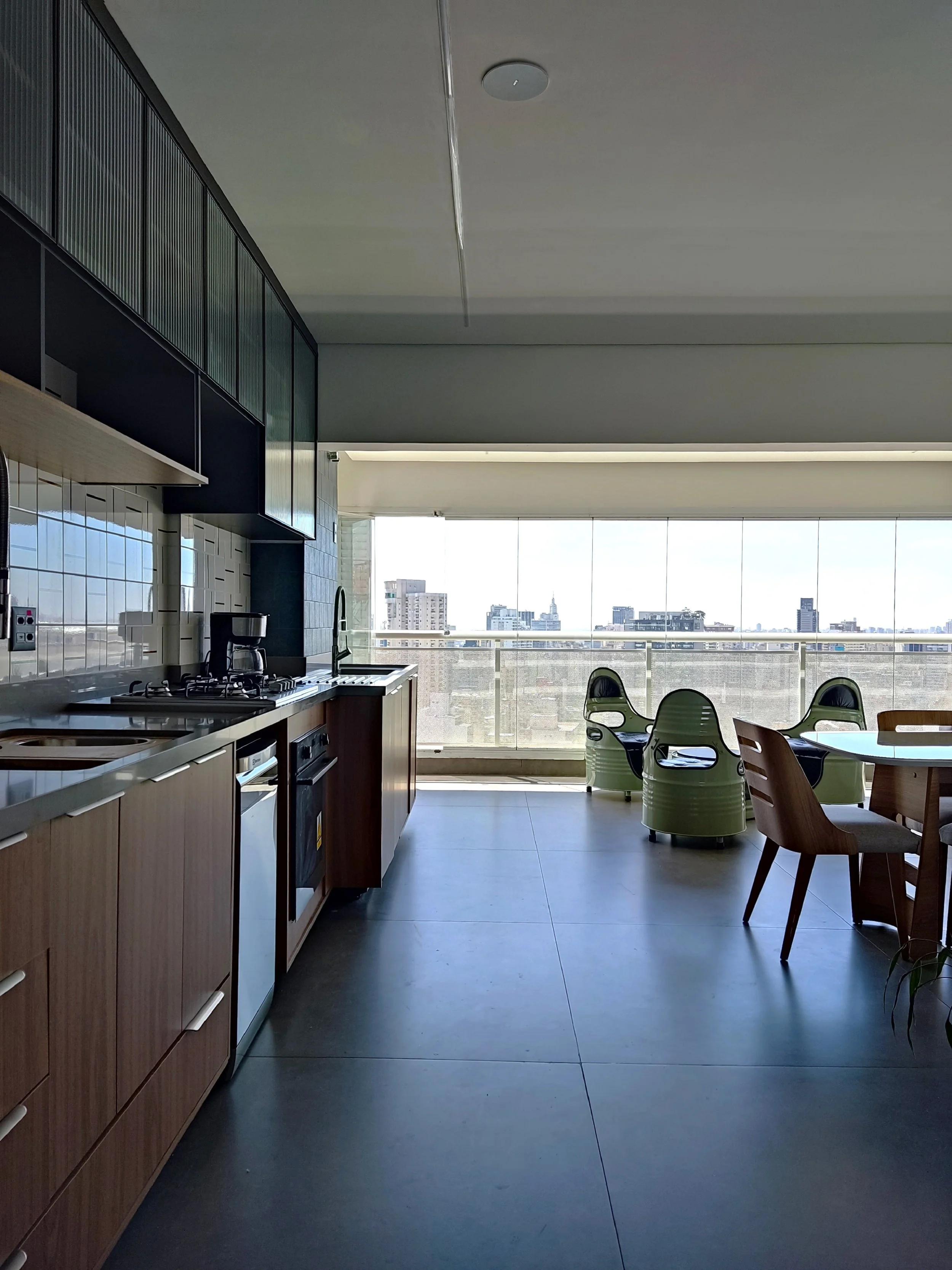 Cozinha moderna com vista para a cidade, com balcão de madeira, armários pretos, pia, fogão e geladeira, além de uma mesa de jantar com cadeiras e poltronas verdes junto à grande janela de vidro