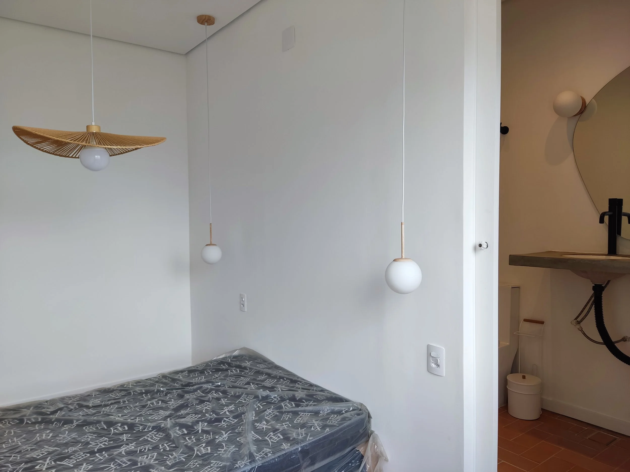 Quarto com cama de colchão novo, parede branca, luminárias pendentes brancas com detalhes de madeira, e banheiro ao lado com pia e espelho arredondado.