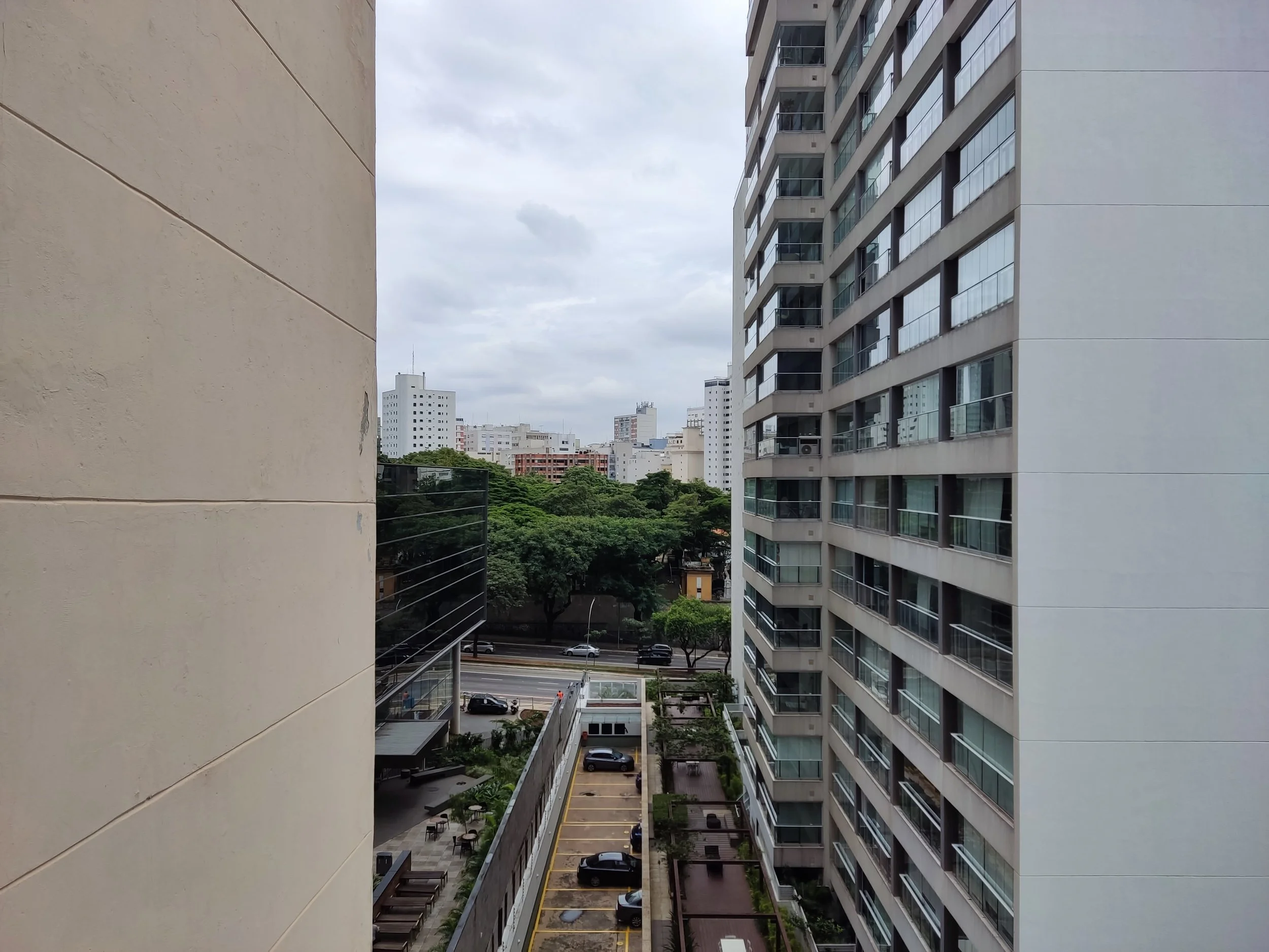 Vista de área urbana com prédios altos, árvores e estacionamento de carros sob céu nublado.