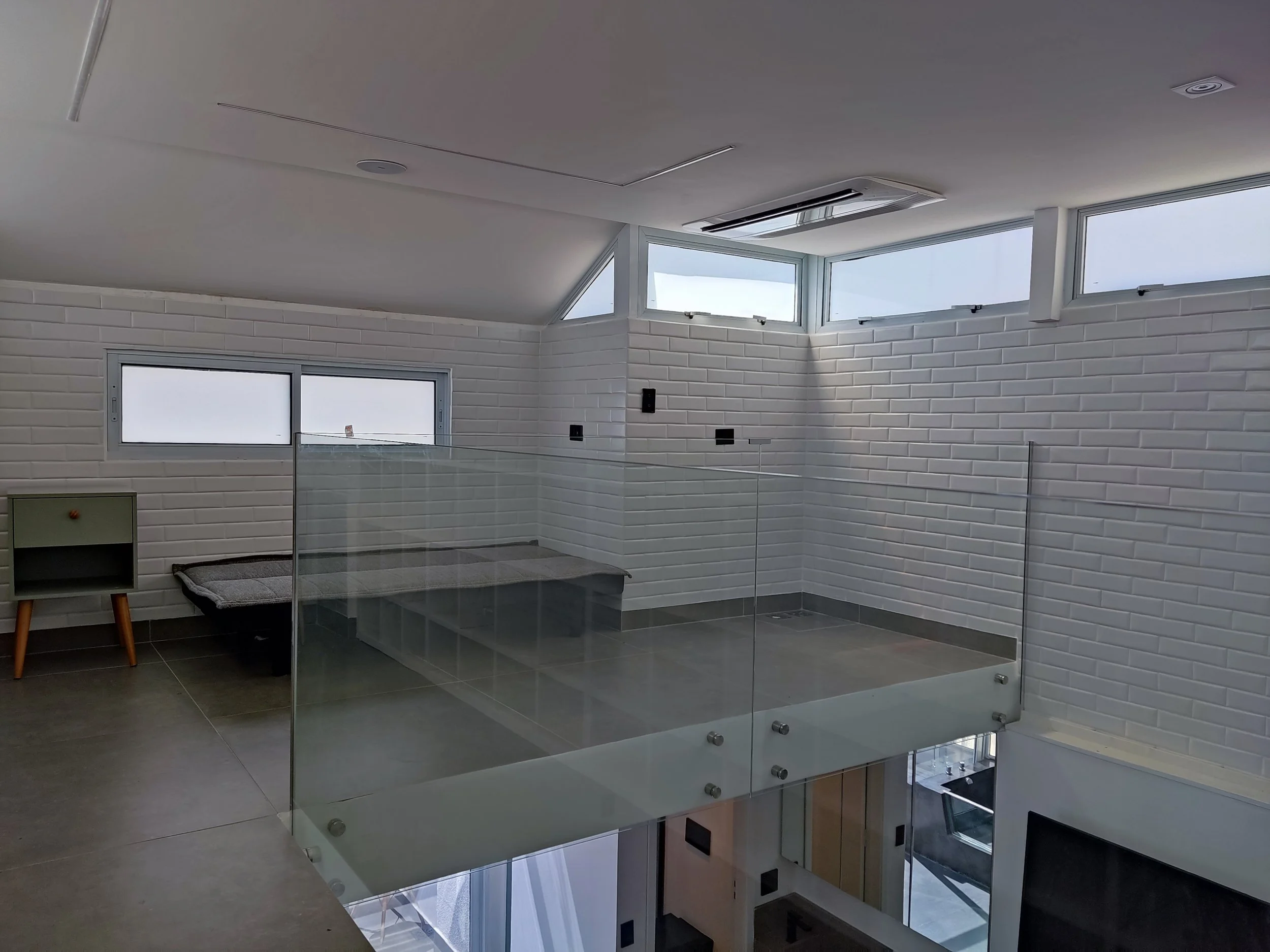 Interior de um quarto com parede de tijolos brancos, janelas altas, teto inclinado e piso de mármore cinza, com uma cama de cachorro e um móvel verde escuro ao lado.