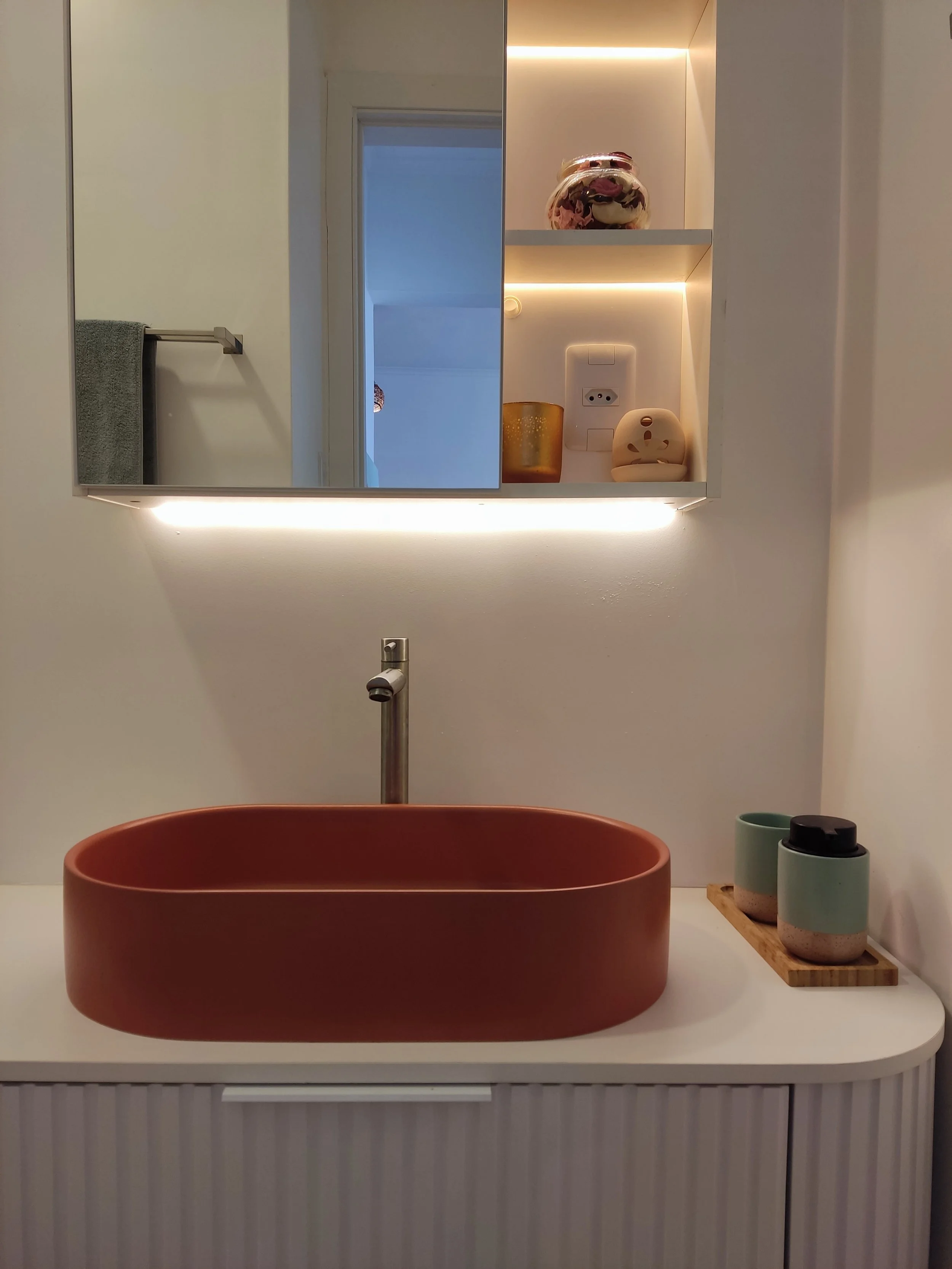 Pia de banheiro com cuba ovalada de cor terracota, espelho com iluminação e prateleira com objetos decorativos e utensílios, incluindo copos e potes
