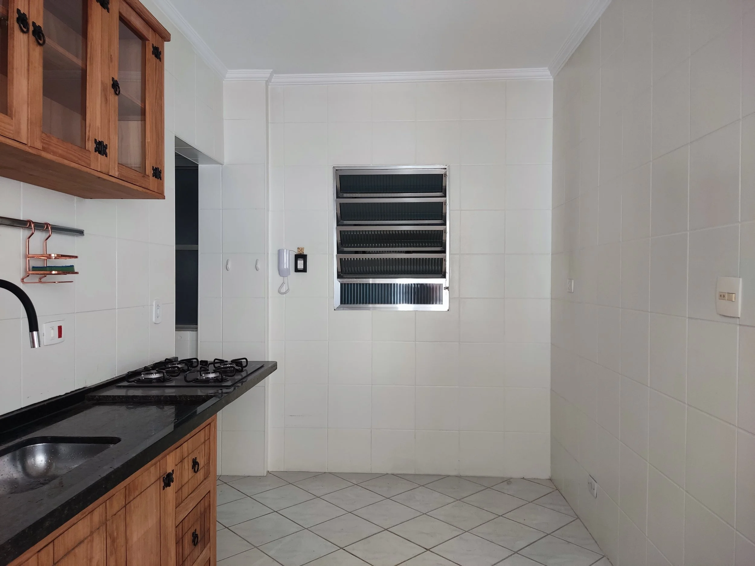 Cozinha com pia de granito preto, armário de madeira, fogão a gás de quatro bocas, janela de vidro com venezianas de alumínio, paredes revestidas de azulejos brancos, piso de azulejos claros, e uma pequena prateleira de cobre na parede do lado esquer