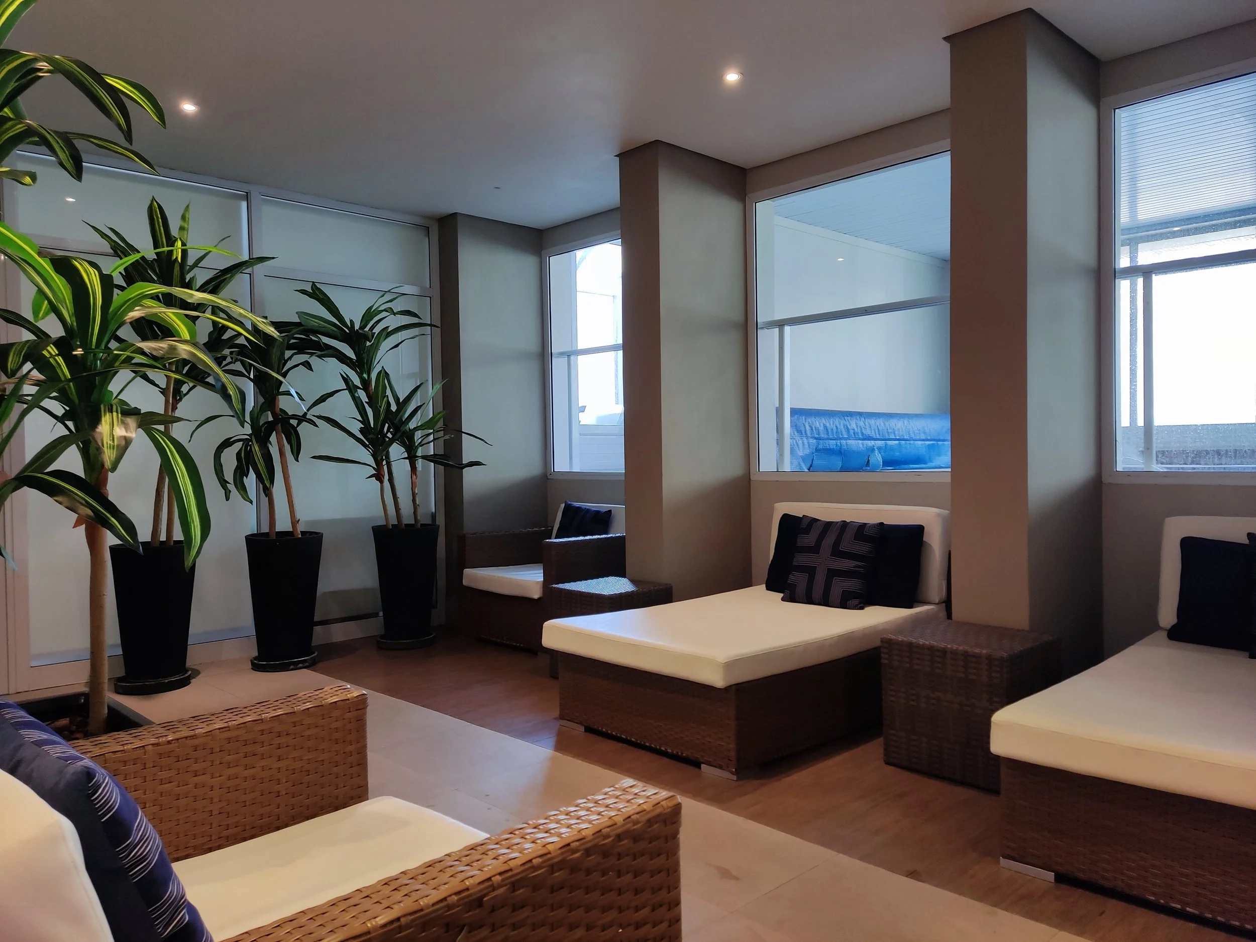 Sala de estar com poltronas de fibra, almofadas preta e branca, plantas altas em vasos pretos e janelas grandes com vista para o exterior.