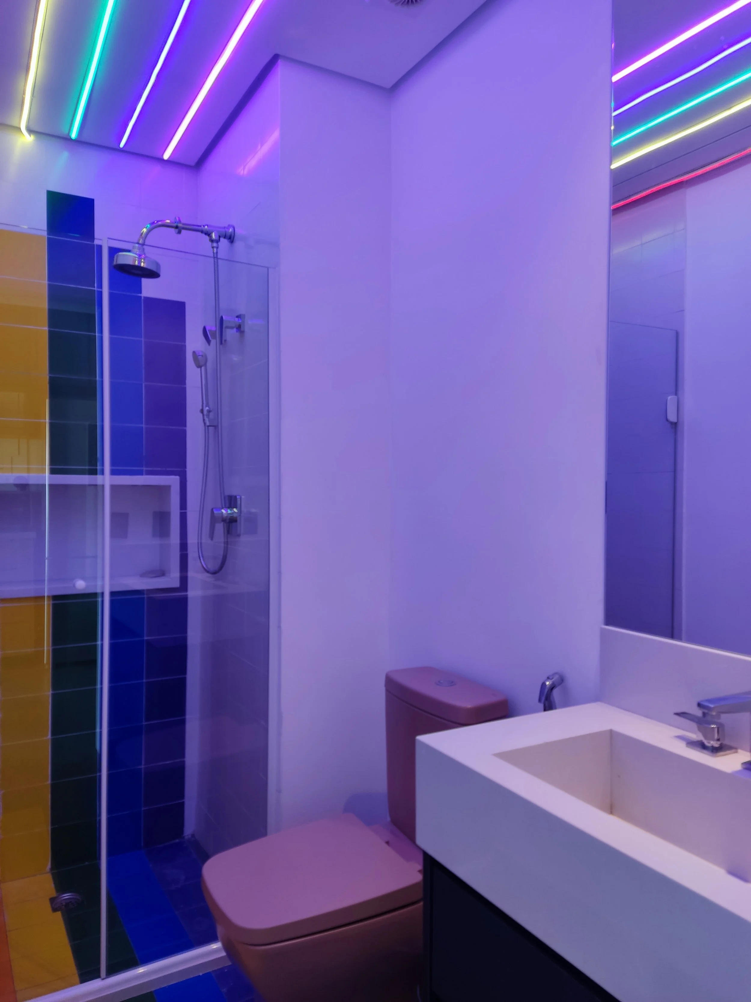 Banheiro com iluminação de LED colorida, ducha, vaso e pia com espelho.