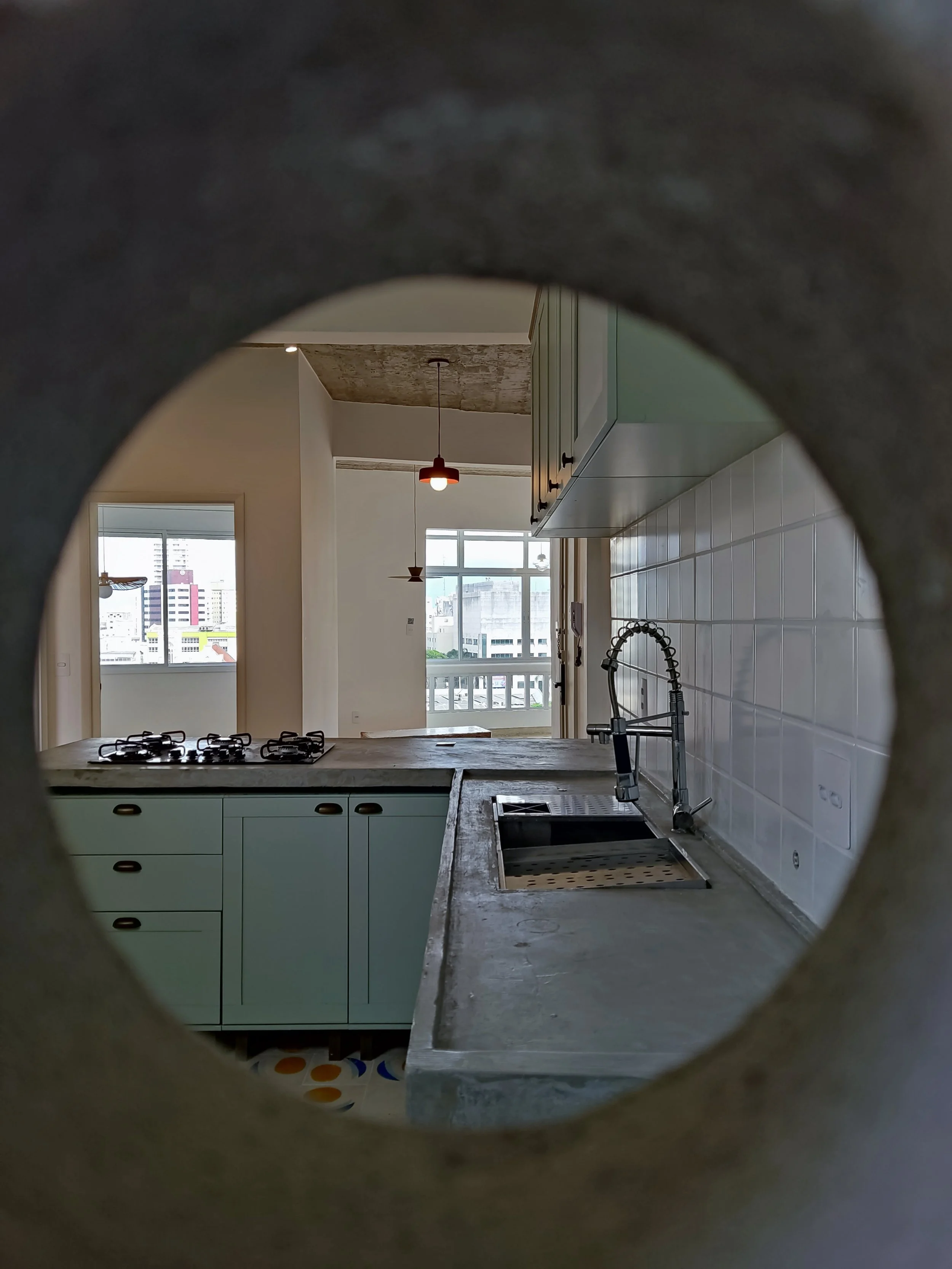 Cozinha moderna vista através de um buraco de uma parede, com armários, pia, torneira de catering, parede com azulejos brancos e janelas que deixam entrar luz natural.