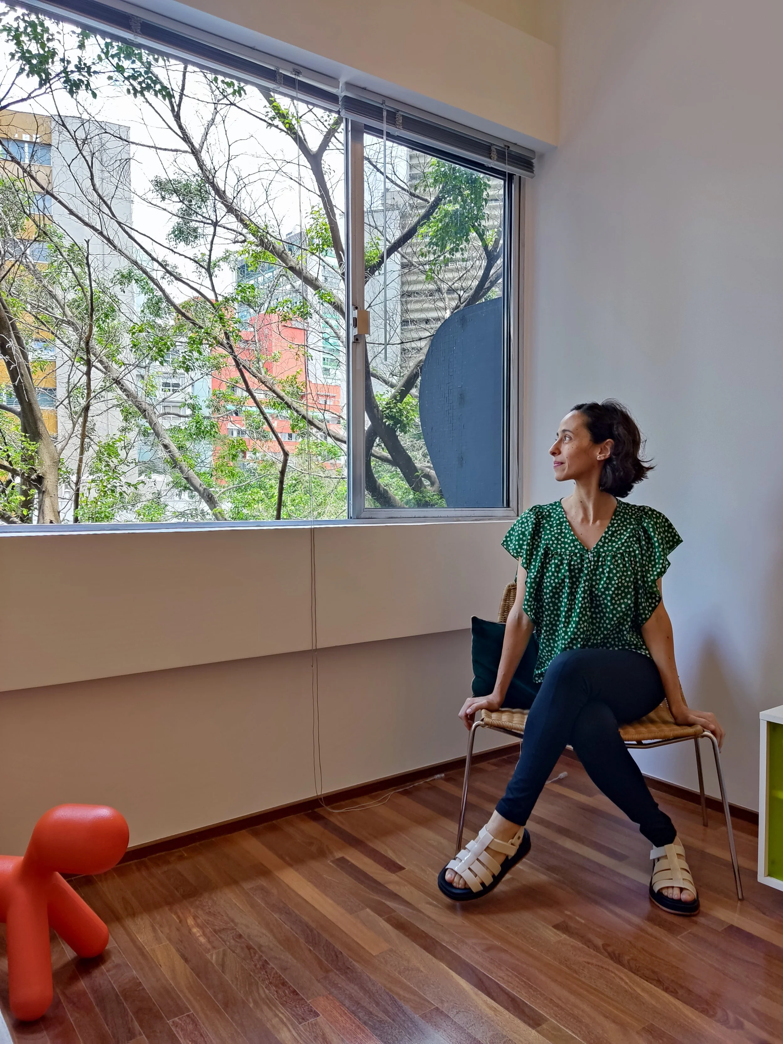 Mulher sentada em uma cadeira perto de uma janela com vista para árvores e prédios na cidade, vestindo blusa verde com bolinhas e sandálias brancas.