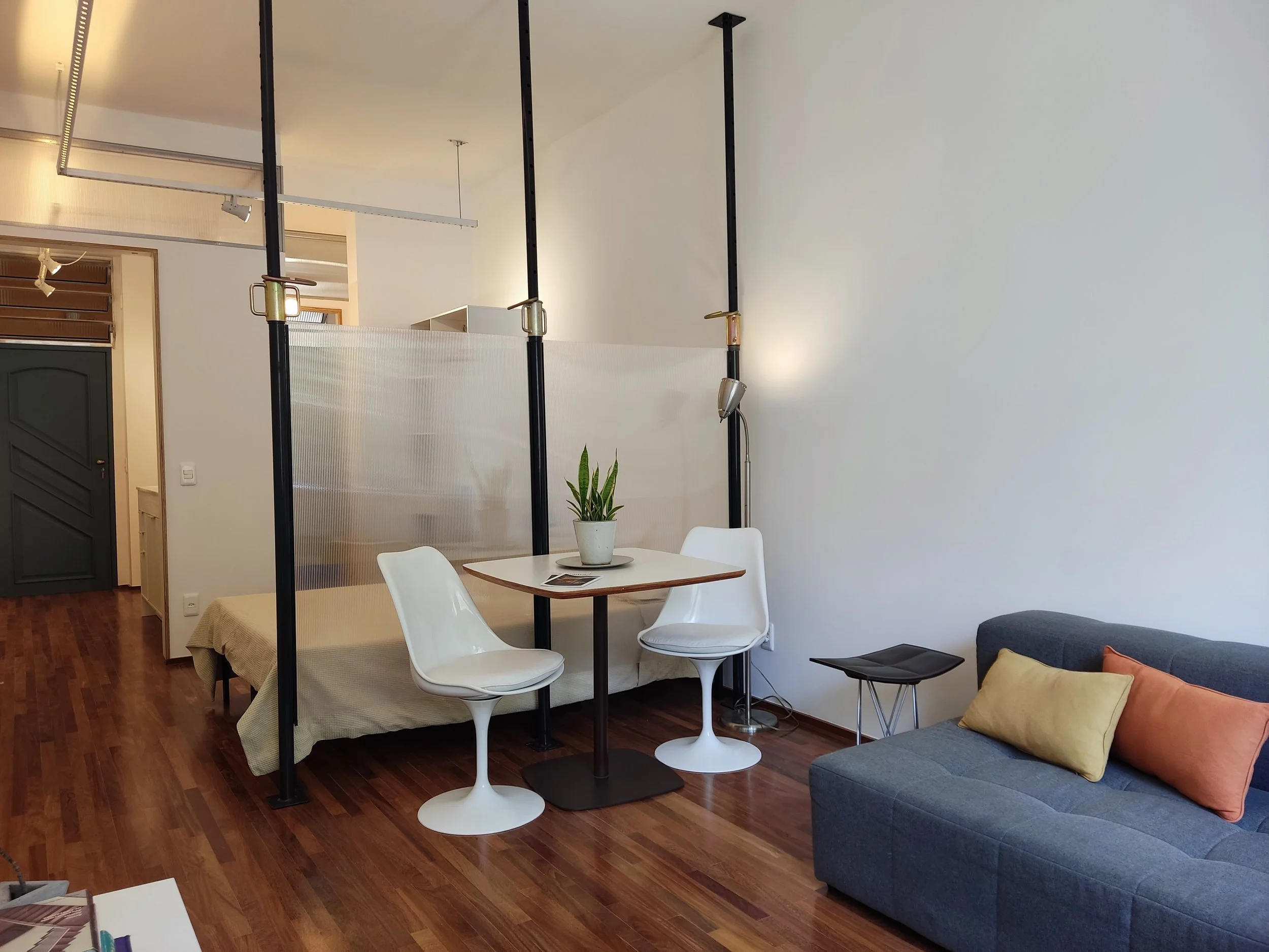 Sala de estar com sofá cinza, almofadas amarela e laranja, mesa de canto preta, mesa de jantar redonda com duas cadeiras brancas, planta no centro da mesa, piso de madeira, divisória de vidro fosco, porta verde na entrada.