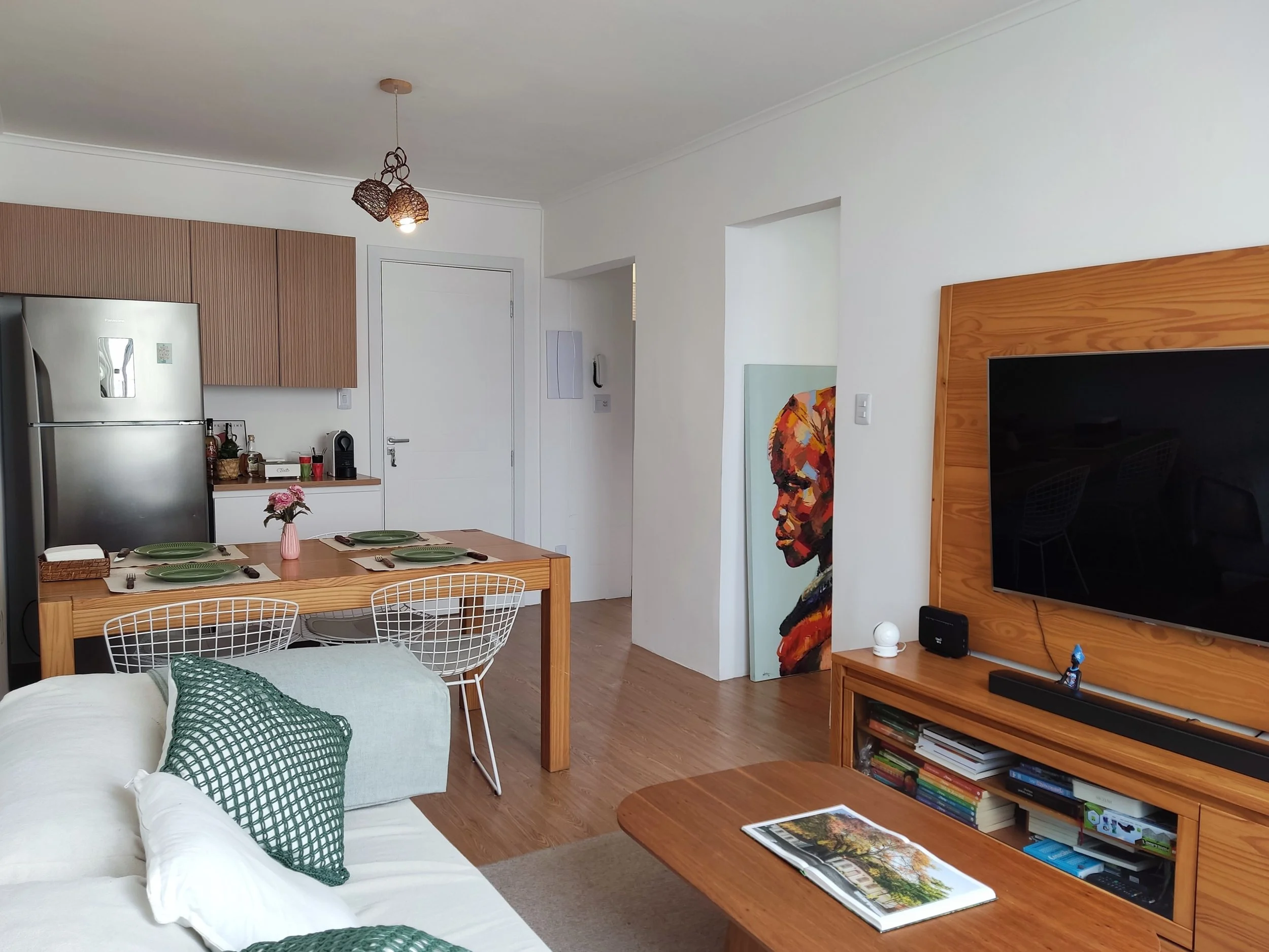 Sala de estar e cozinha com móveis de madeira, televisão, sofá branco com almofadas verdes, mesa de jantar com toalhas, pratos e talheres, quadro de arte e iluminação moderna.