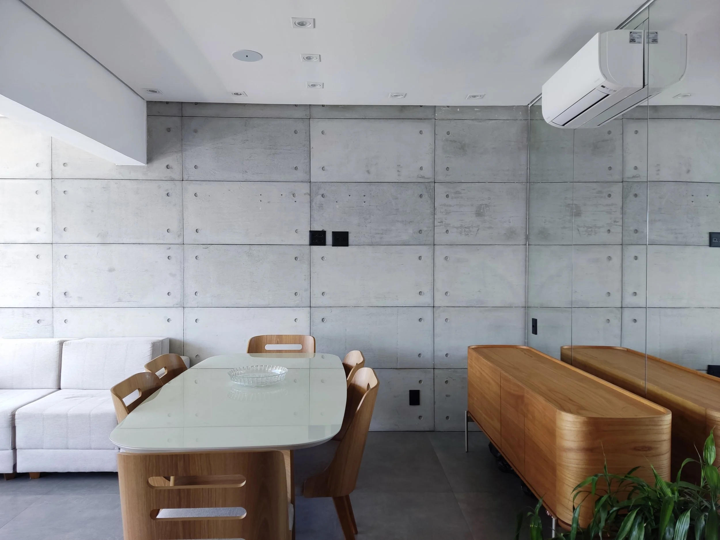 Sala de jantar moderna com parede de concreto, mesa de vidro branca, cadeiras de madeira, um sofá branco, um móvel de madeira e ar-condicionado no teto.