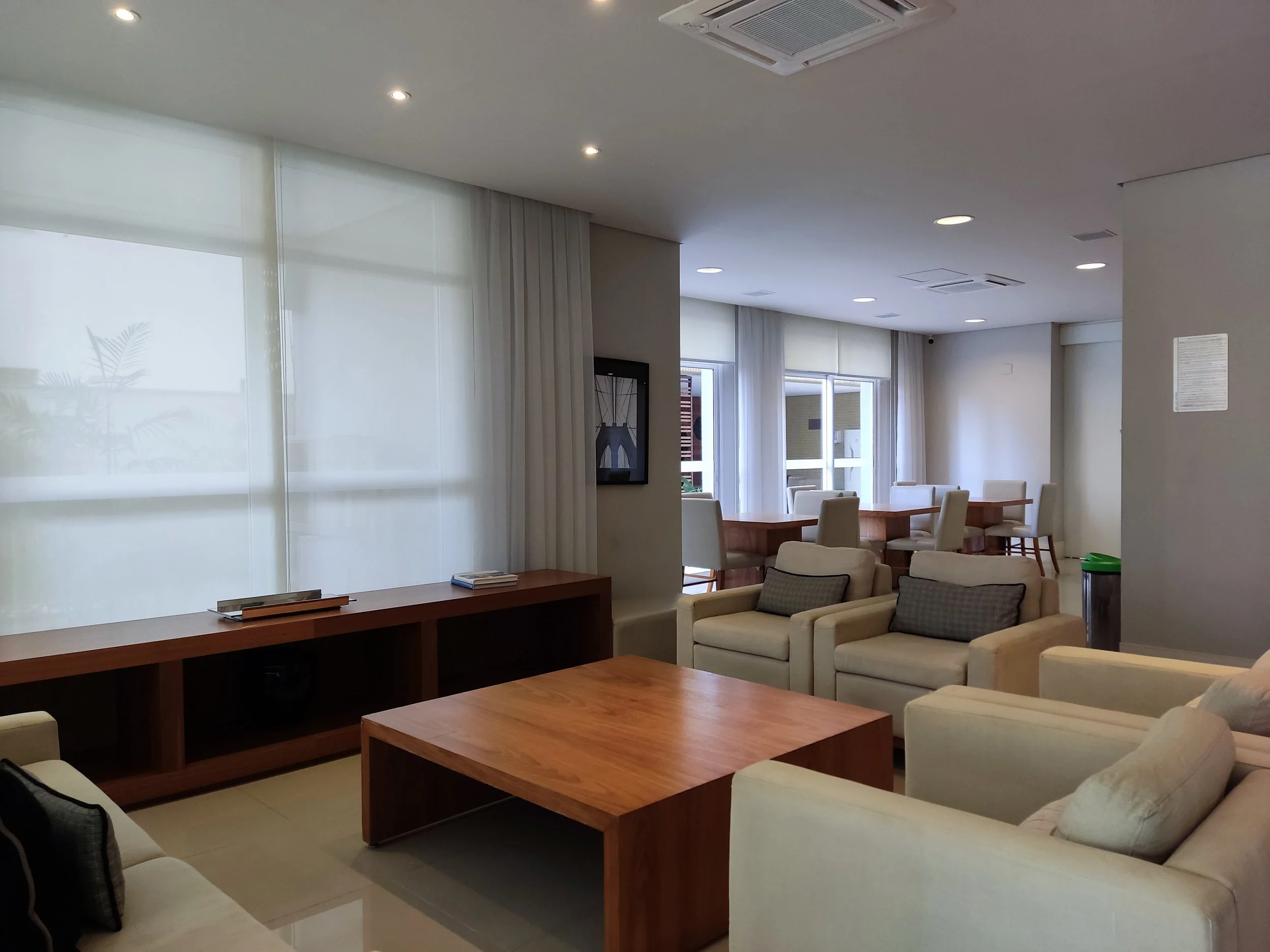 Sala de estar com sofá, poltronas, mesa de centro e uma mesa de jantar ao fundo, com grandes janelas com cortinas brancas.