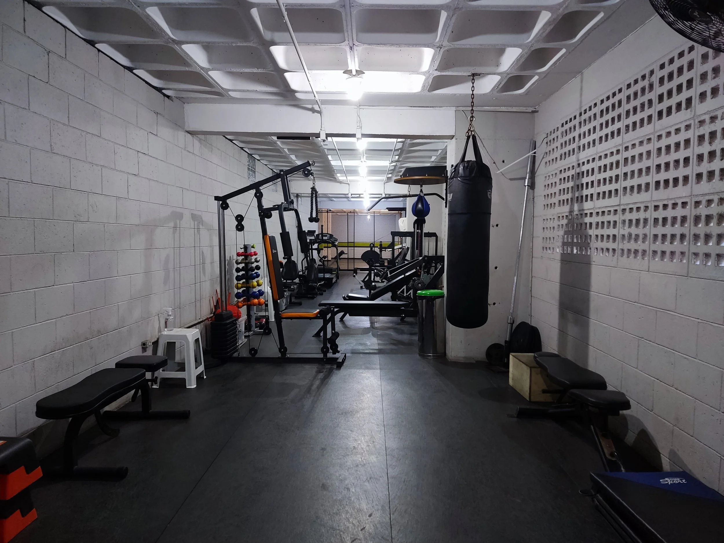 Ginásio com equipamentos de treino, como banco de supino, saco de pancadas, pesos, e vários aparelhos de musculação, com piso de borracha e paredes de tijolos.