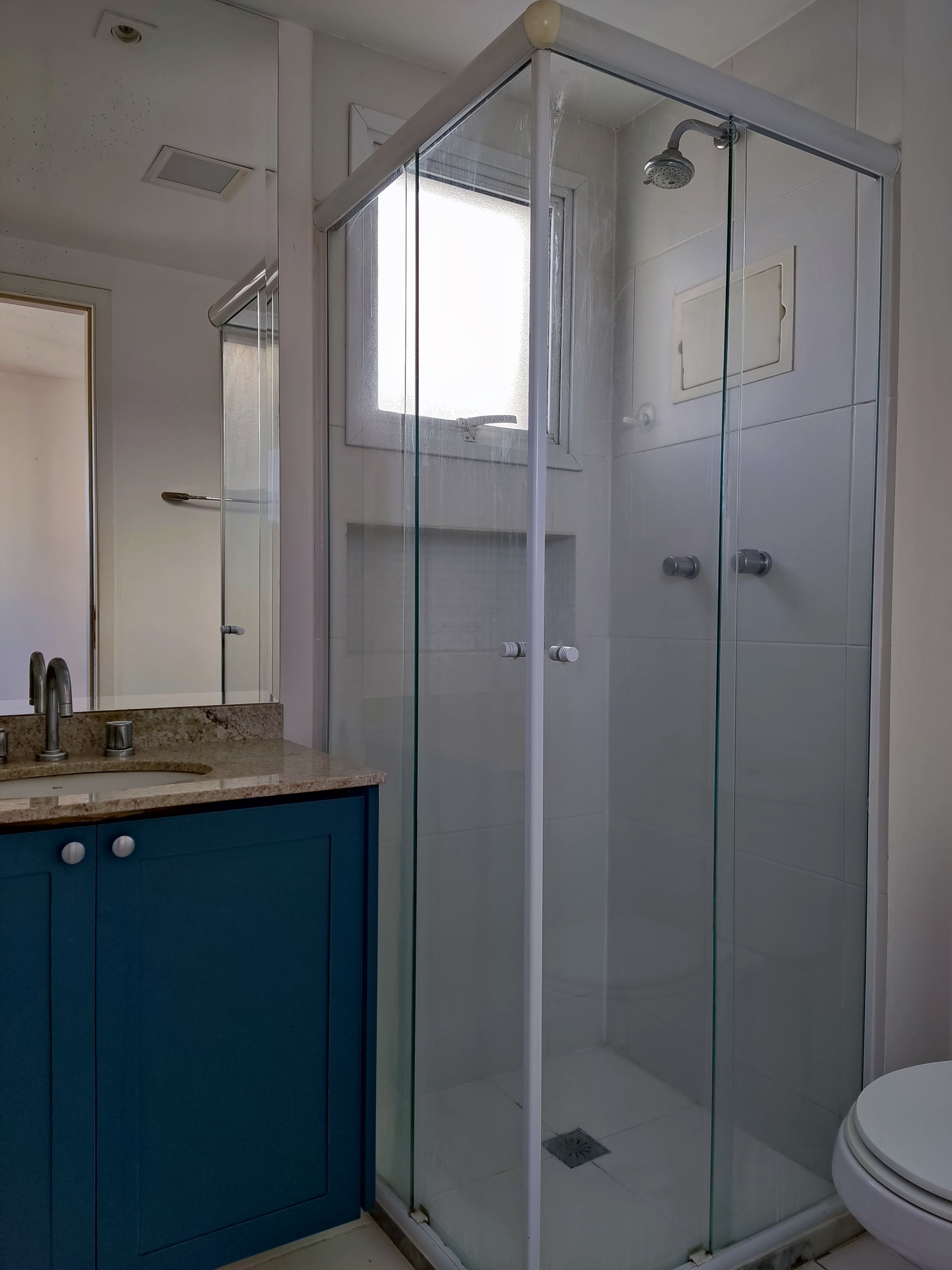 Banheiro com pia de granito azul, espelho grande, box de vidro com ducha, janela, vaso sanitário parcialmente visível, paredes brancas e piso de azulejos.