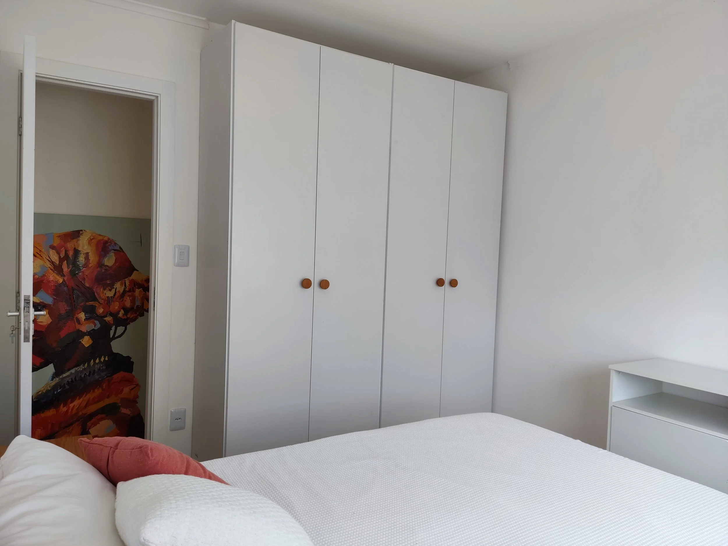 Quarto com cama de casal, armário branco com portas de madeira de pua, gaveta branca com prateleira, parede branca, porta entreaberta com mural de caveira de cores quentes ao fundo.