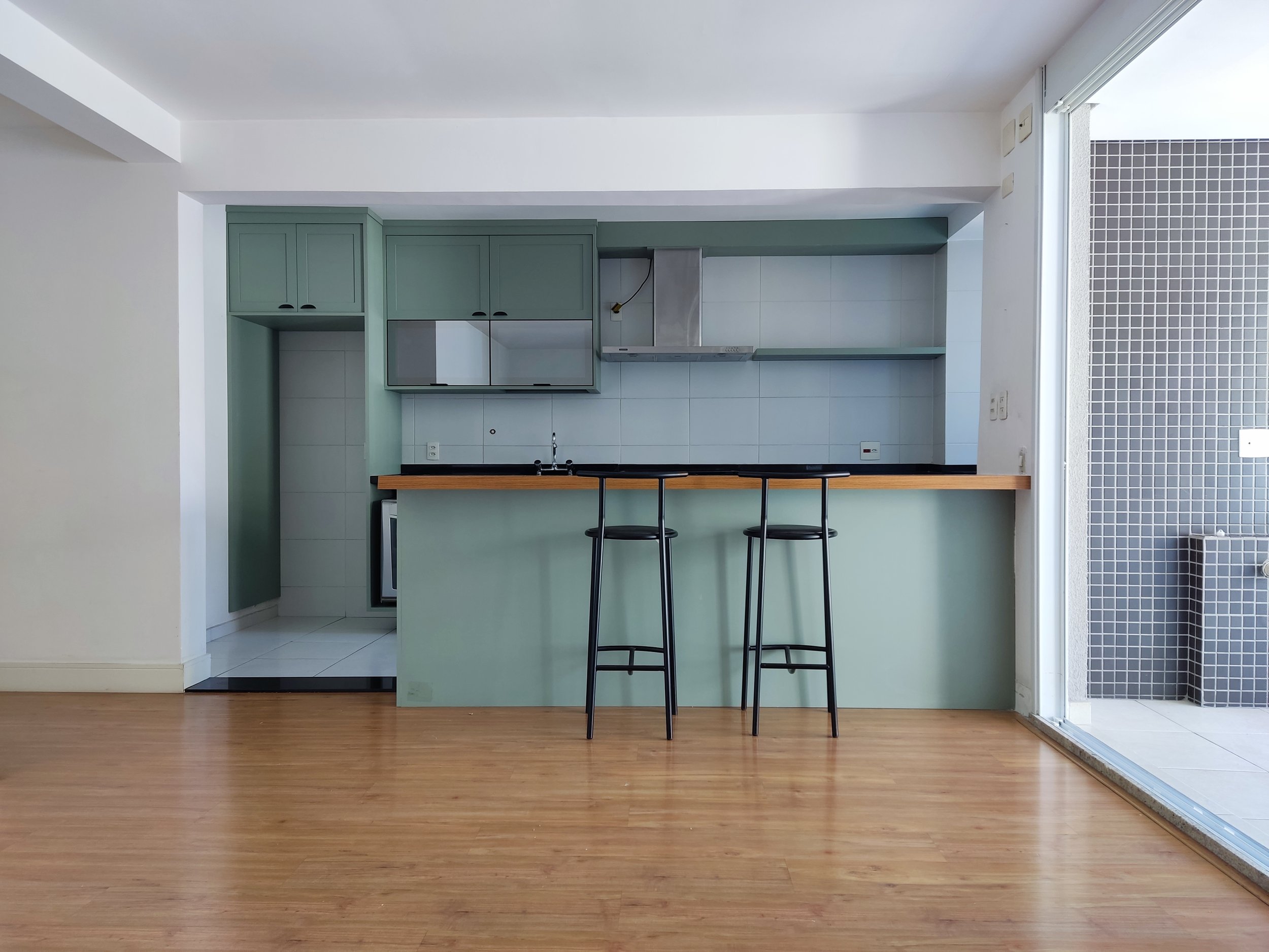 Cozinha moderna com teto branco, armários verdes, bancada de madeira, janelas de vidro, piso de madeira e uma área de lavanderia com azulejos escuros ao lado.