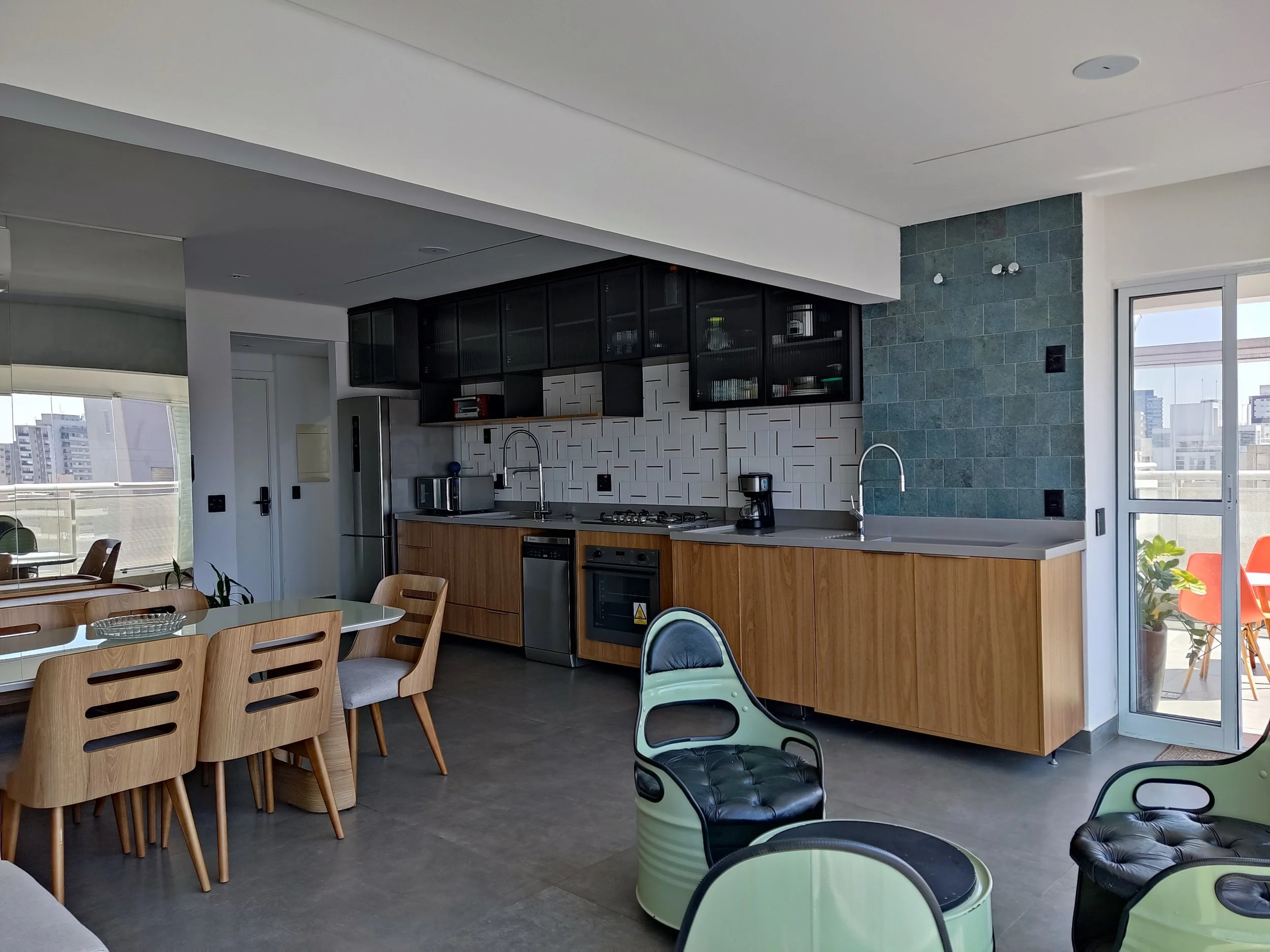 Sala de jantar e cozinha moderna com móveis de madeira clara, balcão de cozinha de madeira, armários pretos, eletrodomésticos de aço inox, paredes com revestimento branco e azul, grande janela com varanda e vista da cidade.