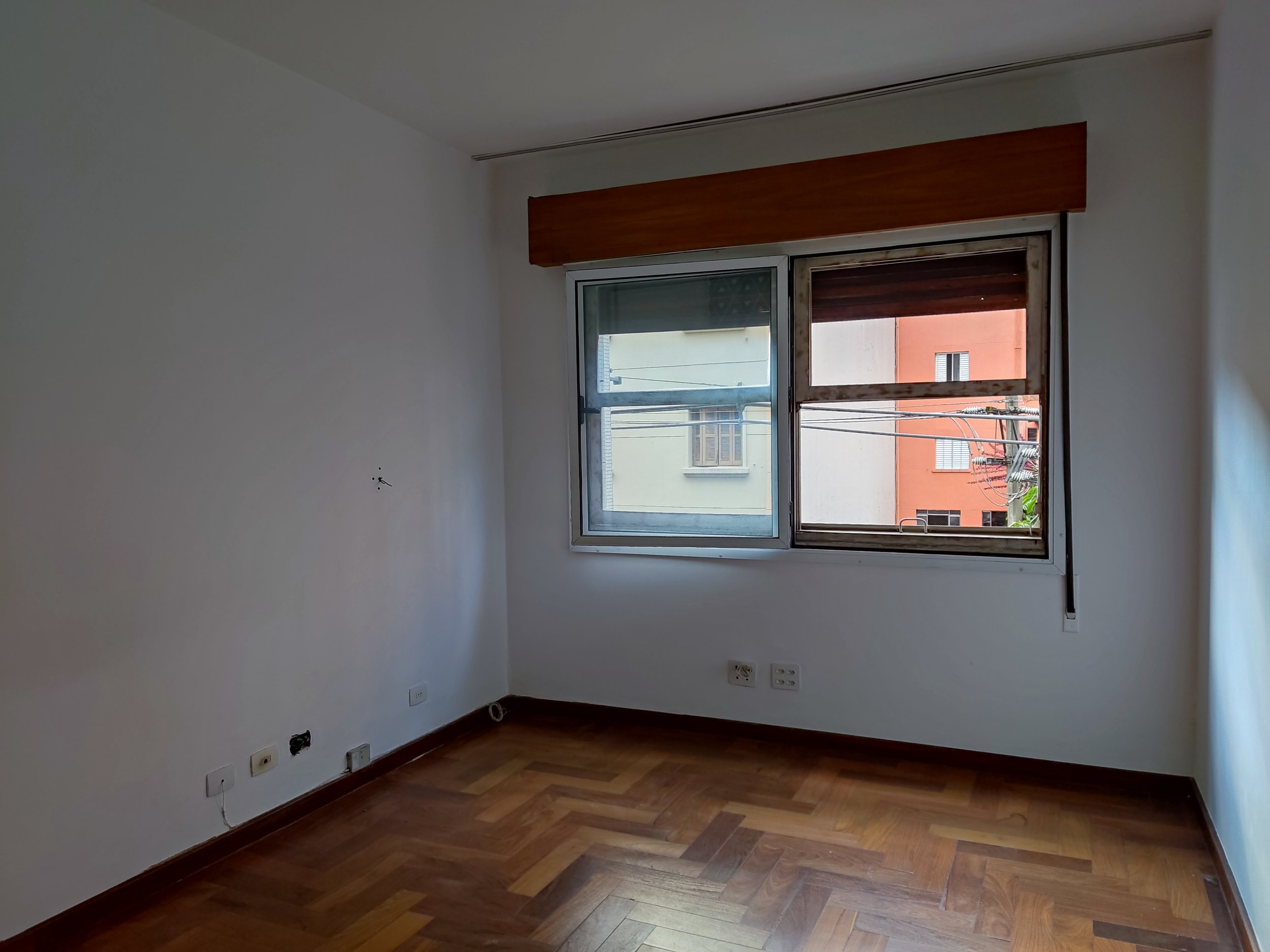 Sala vazia com piso de madeira, uma janela com duas folhas - uma de vidro e uma de ferro velho, na parede branca. A janela mostra edifícios residenciais ao lado, parede de tijolos e fios elétricos.