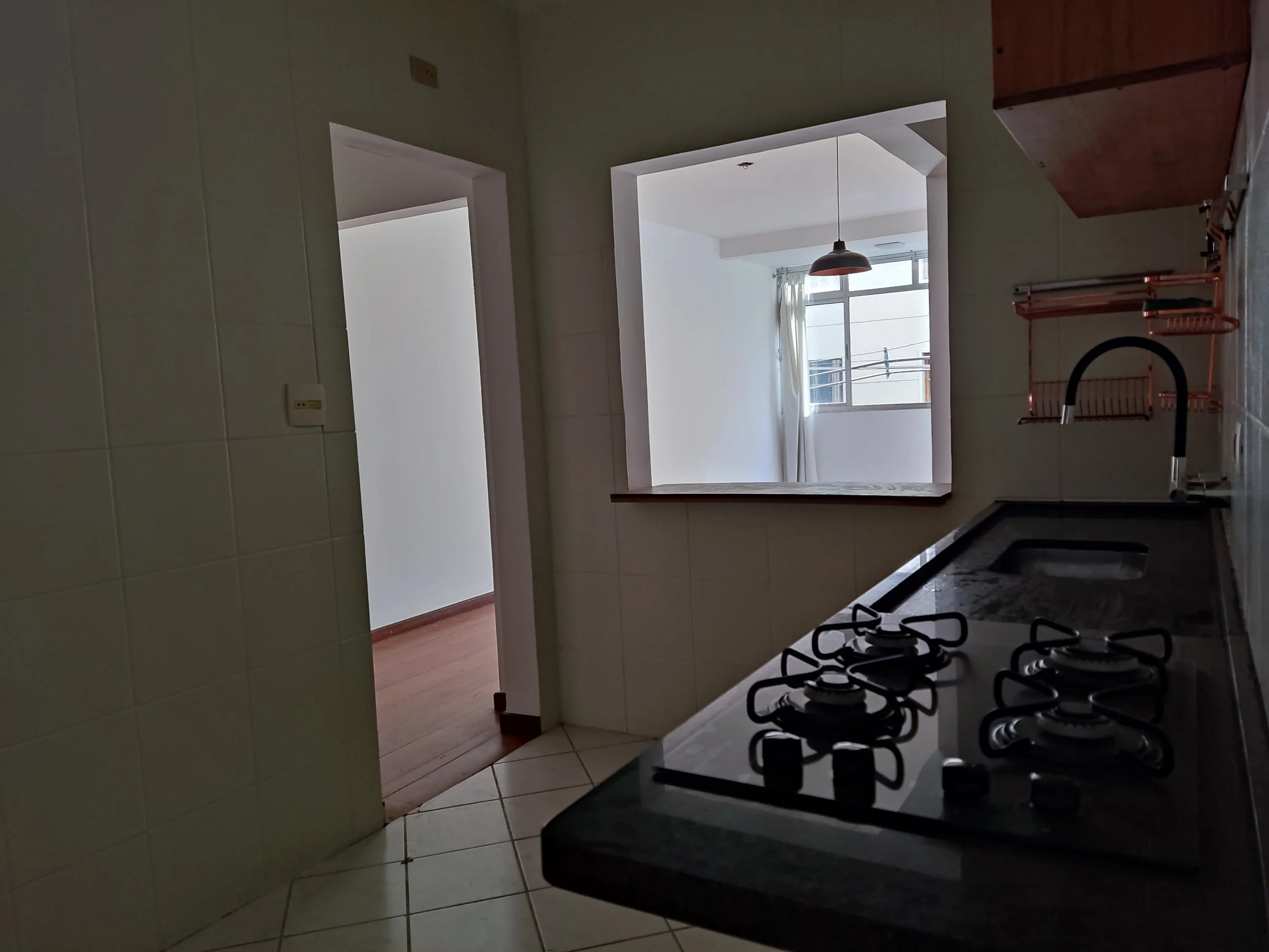 Cozinha com pia de ferro e fogão a gás na parede, parede com revestimento branco, porta que leva a outro cômodo, janela com cortina branca e vista de prédios externos