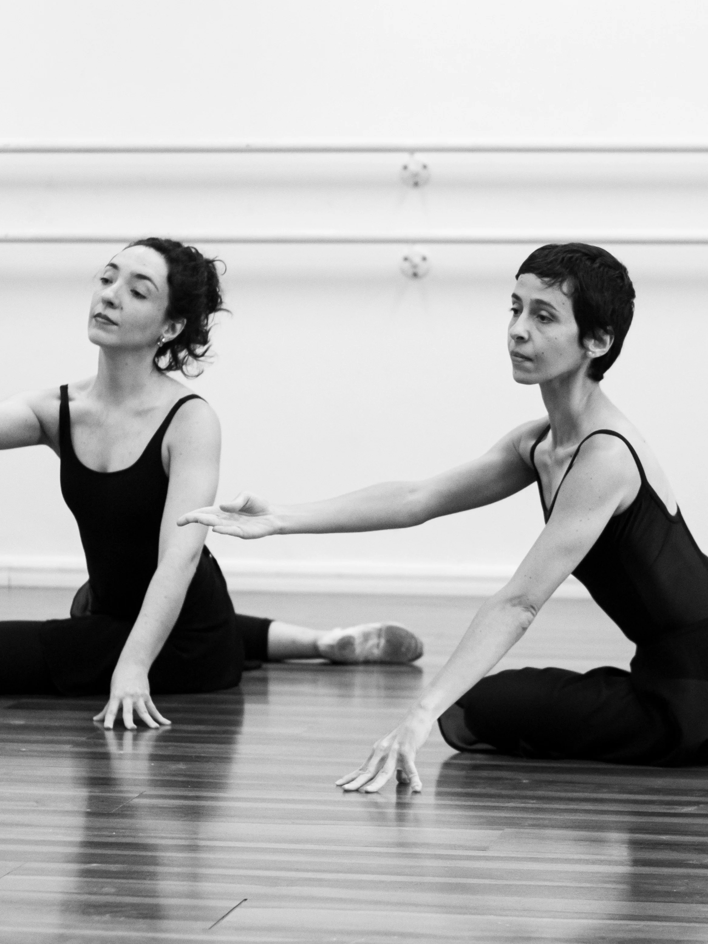 Duas mulheres dançarinas de ballet praticando alongamento no chão de madeira, em um estúdio de dança. Ambas usam roupas pretas e estão em posição de alongamento, uma com o braço estendido para alcançar o chão e a outra com a cabeça levemente inclinada, em um ambiente de prática de ballet.