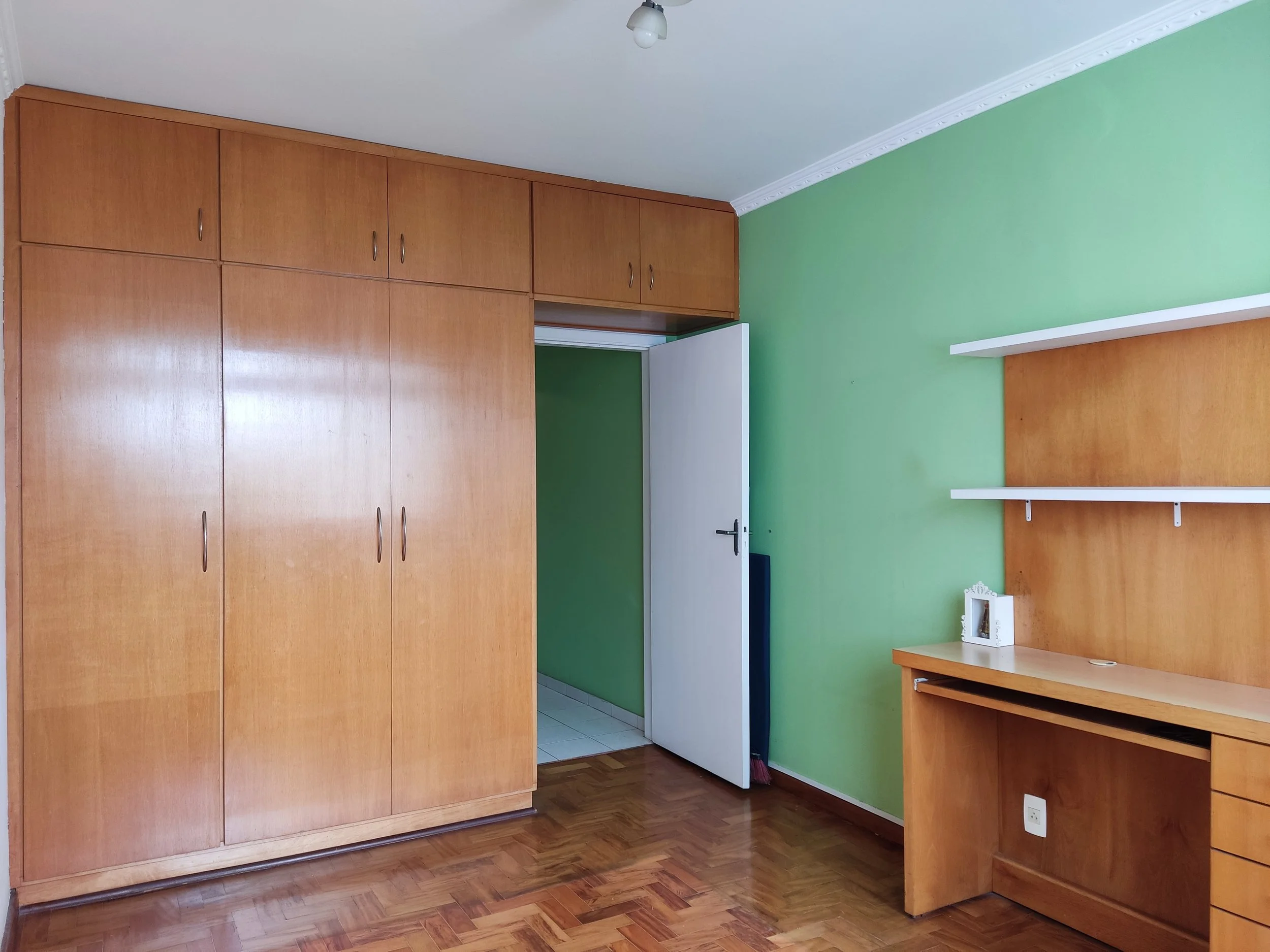 Quarto com parede verde, armário de madeira, escrivaninha de madeira com prateleiras brancas, chão de parquet, porta branca aberta, objetos decorativos e tomada elétrica.
