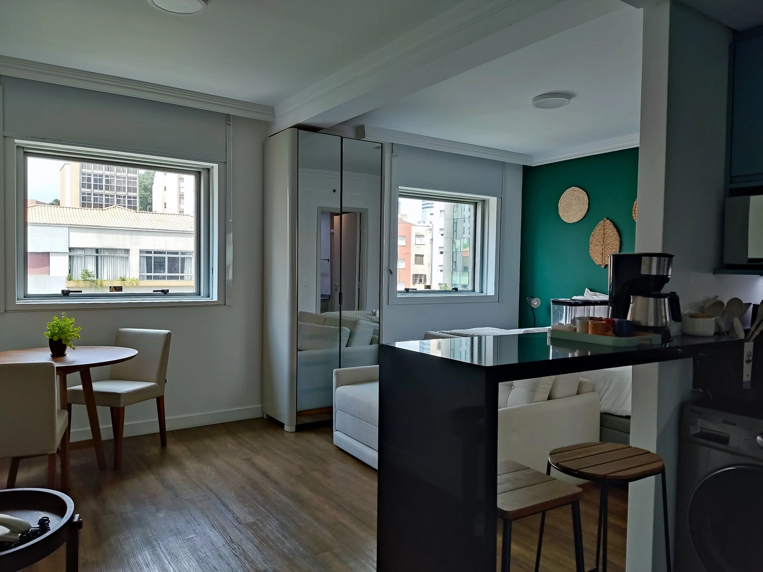 Sala de estar e cozinha com janelas grandes, parede verde com decoração de cestas de palha, mobília branca, mesa redonda com cadeira e cozinha com bancada preta, bebidas e utensílios.