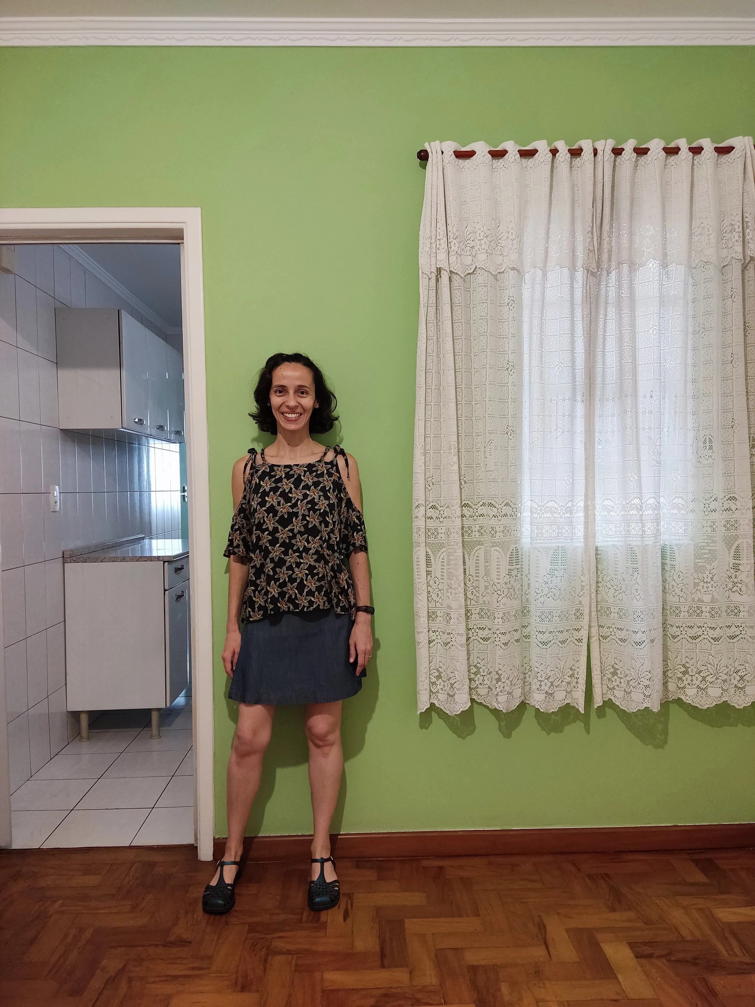Mulher sorridente de cabelo escuro, usando blusa com alças, saia curta de jeans e sapatos pretos, posando dentro de casa ao lado de uma parede verde e uma janela com cortinas brancas.