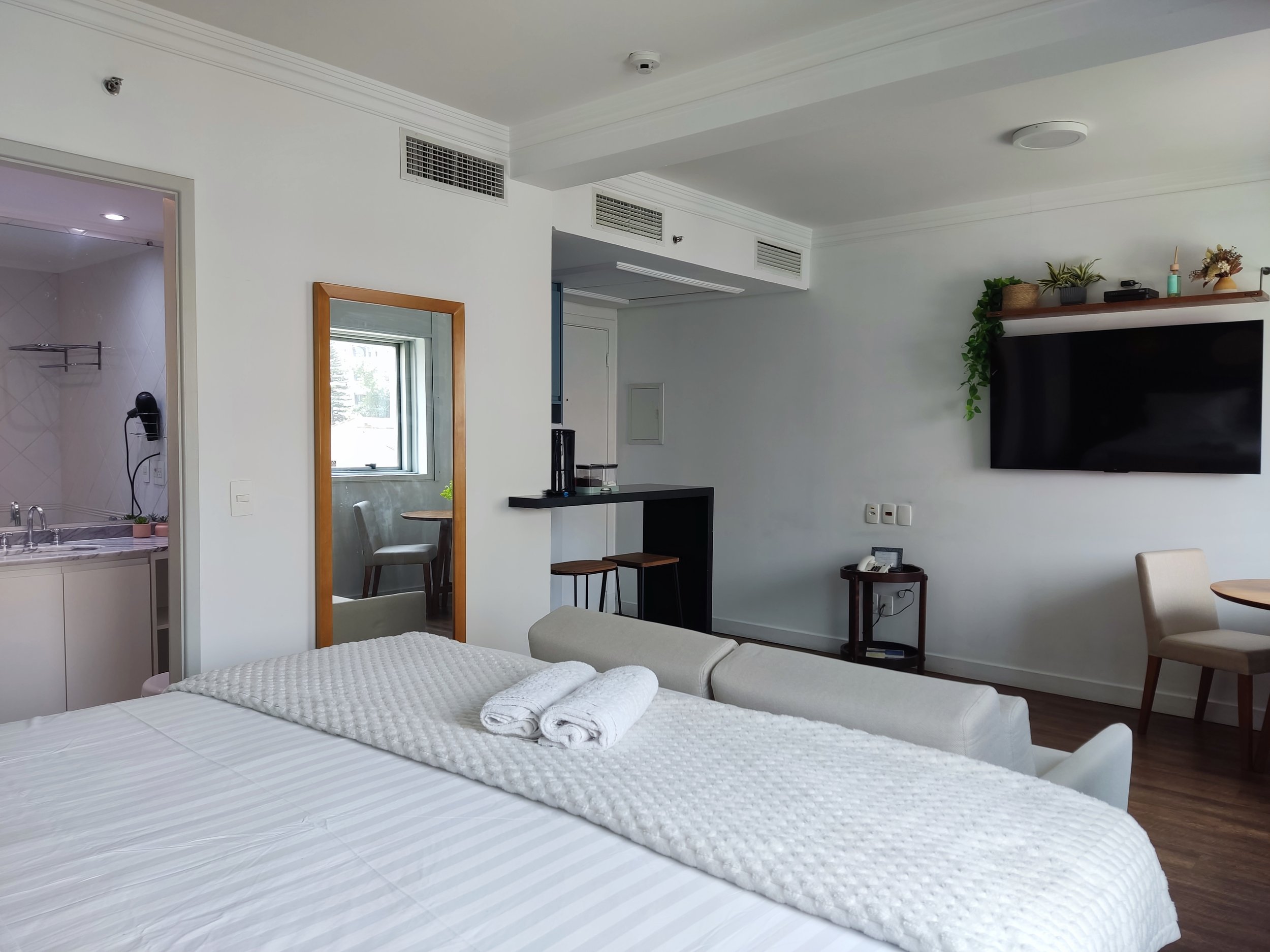 Quarto de hotel com cama de solteiro, toalhas dobradas, espelho de corpo inteiro, TV de tela plana na parede, bancada com café, cadeira, mesa de madeira, parede branca, decoração com plantas e objetos decorativos.
