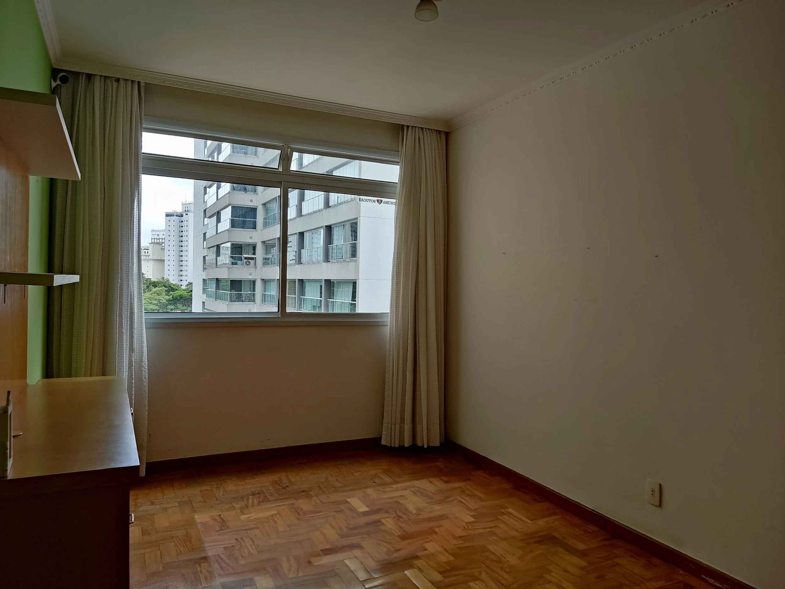 Quarto vazio com janela grande, cortinas claras, piso de madeira e paredes claras.