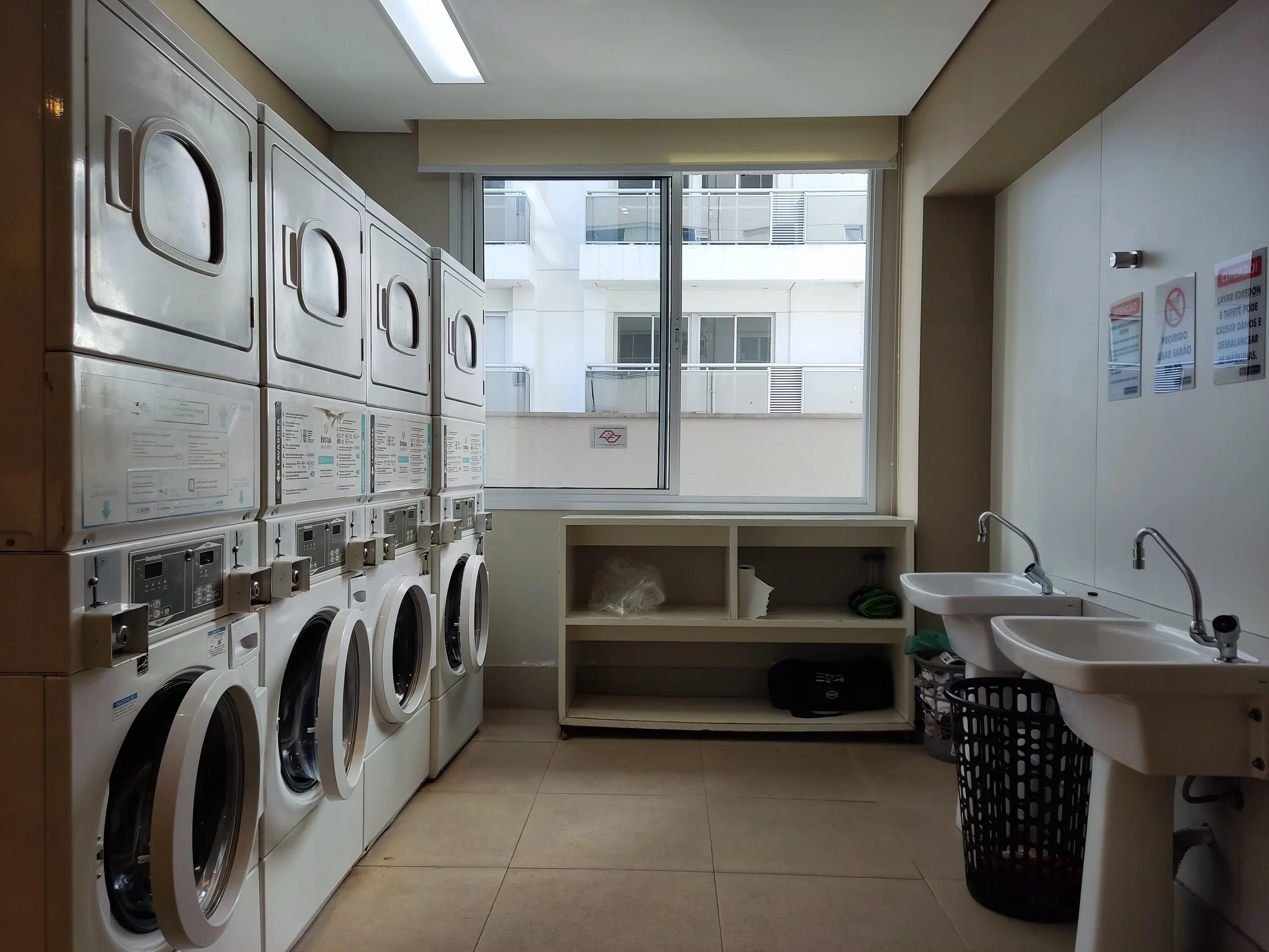 Lavanderia comunitária com máquinas de lavar e secar, duas pias, prateleira vazia, janela grande e paredes brancas.