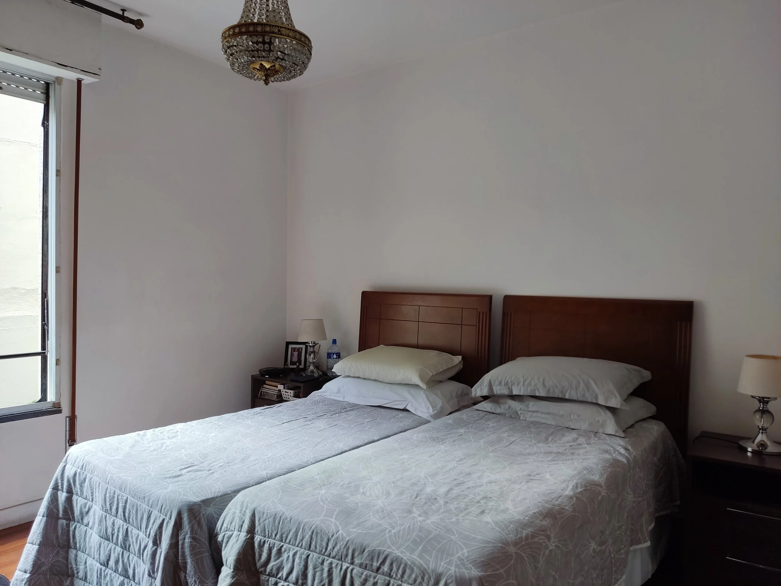 Quarto com duas camas de solteiro com cabeçalhos de madeira, uma de lado da outra, com travesseiros brancos, uma de uma cor clara e a outra branca, e edredons brancos com detalhes em relevo. Ao lado, há duas mesas de cabeceira com abajures, um com um