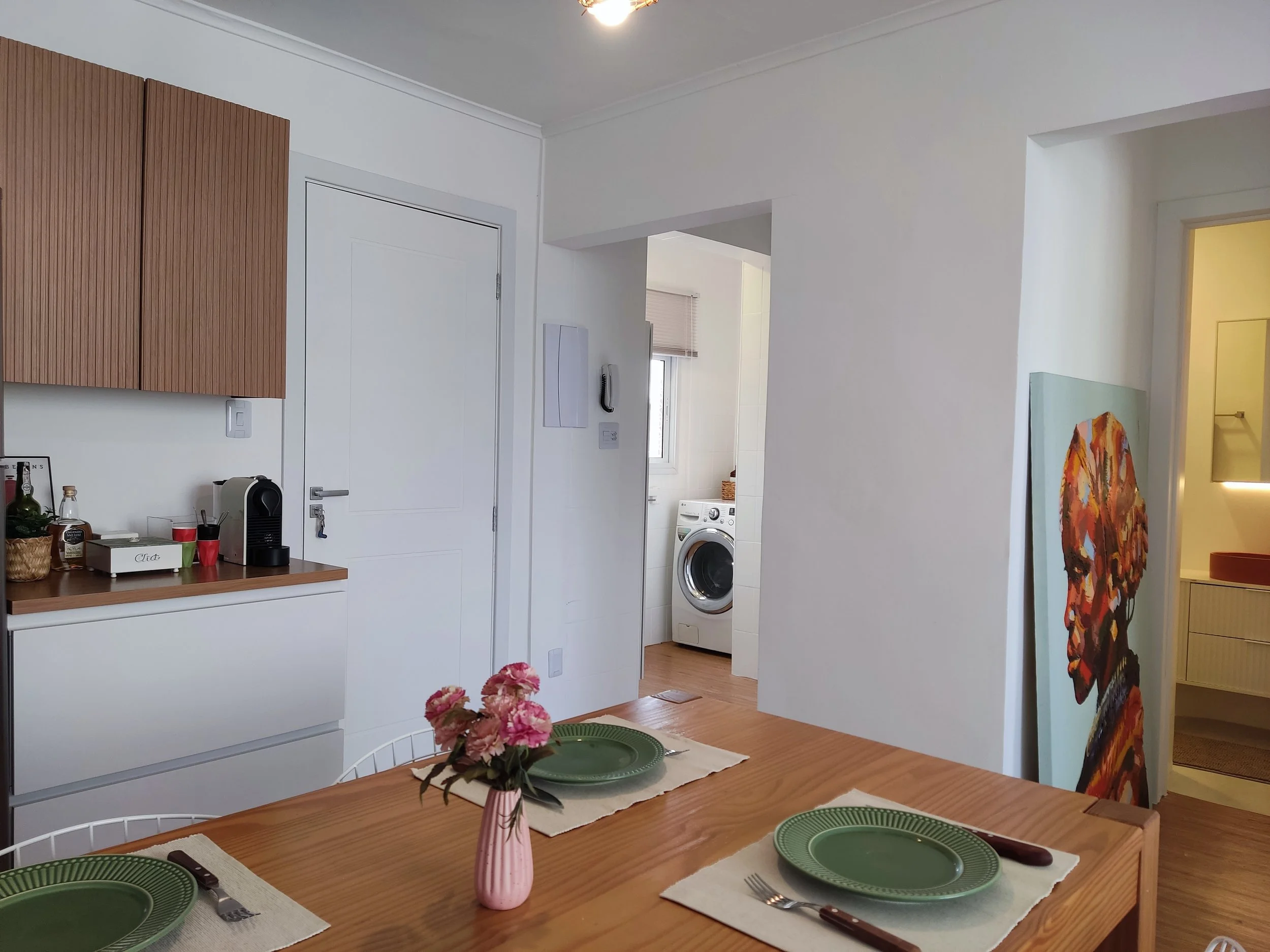 Sala de jantar com mesa de madeira, quatro cubiertos, pratos verdes, um vaso com rosas, cozinha ao fundo com armários brancos e uma porta que leva a uma lavanderia com máquina de lavar.