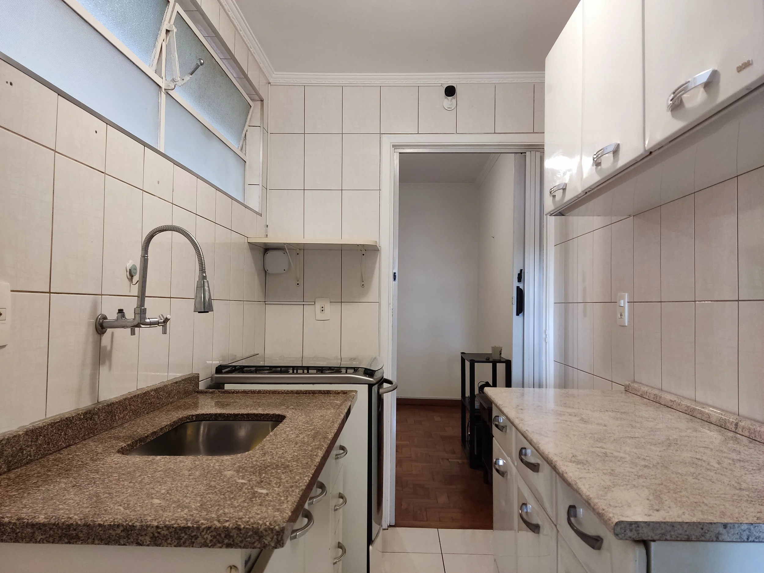 Cozinha com bancadas de granito, pia, fogão, armários brancos, parede com azulejos brancos, janela com vidro fosco, porta de correr levando a outro ambiente com piso de madeira.