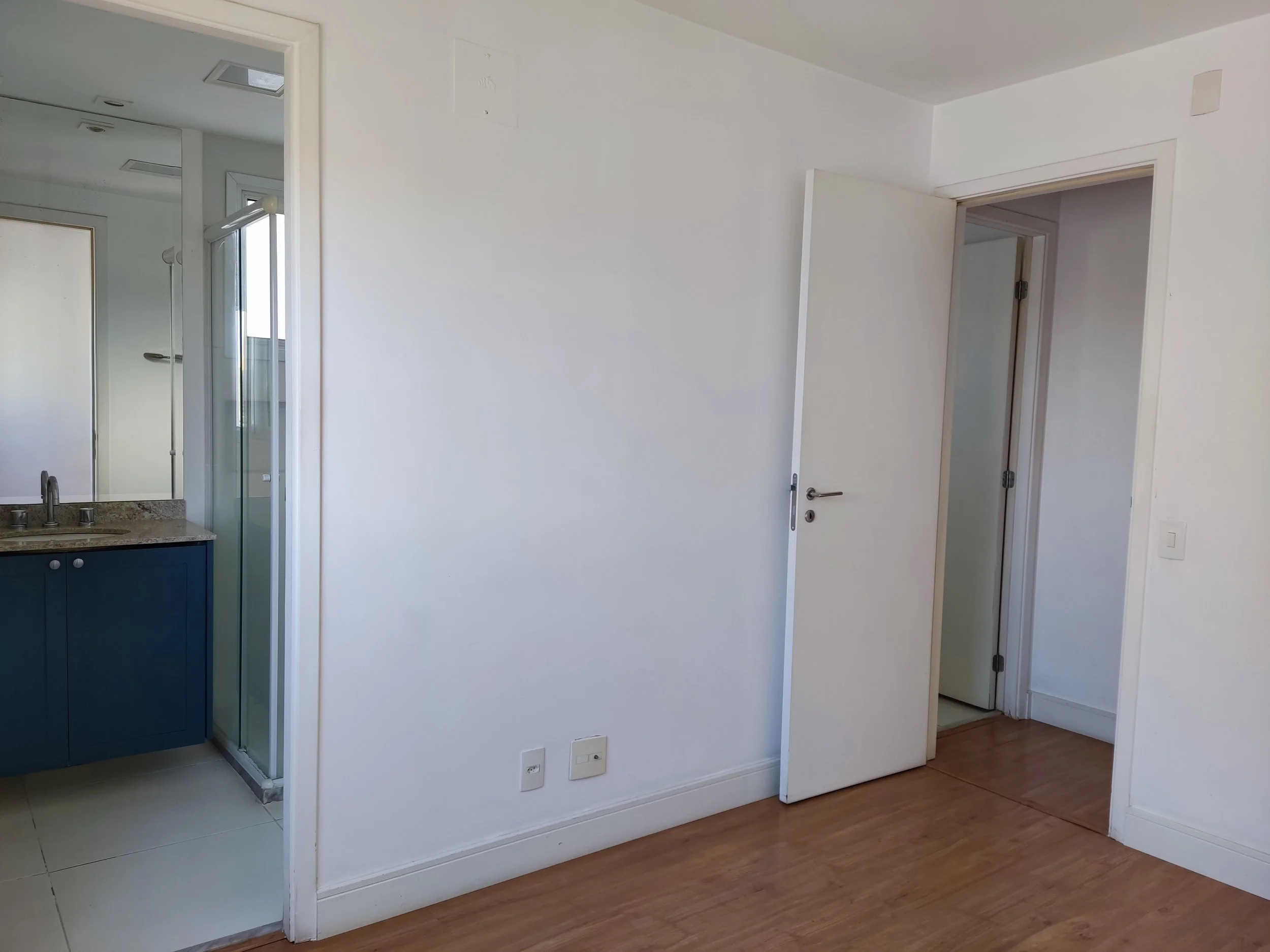 Quarto vazio com piso de madeira, porta aberta para o banheiro, e uma porta fechada ao lado da parede.