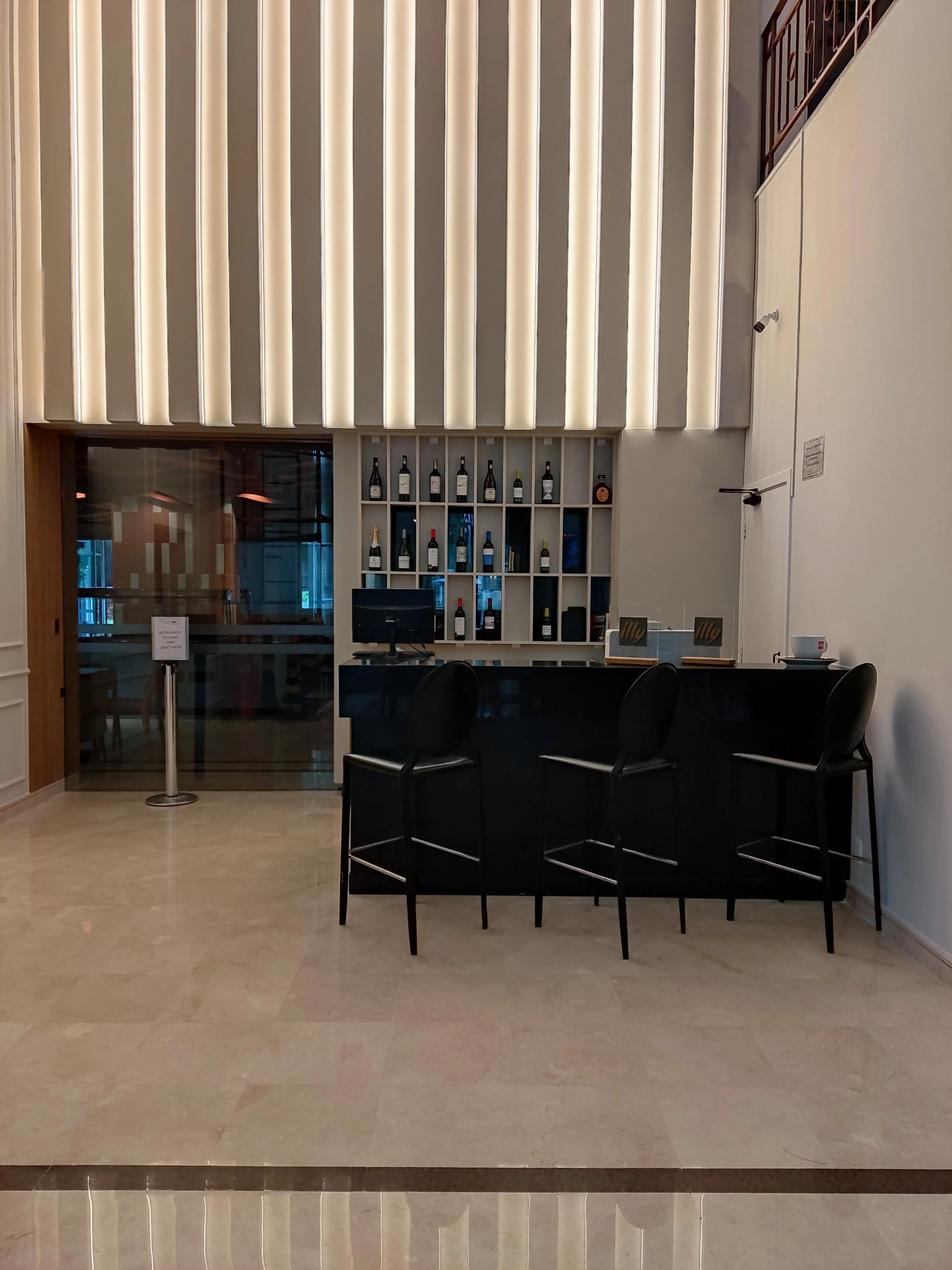 Bar de hotel com balcão preto, três banquetas pretas, parede com prateleira de garrafas de vinho e porta de vidro ao fundo.