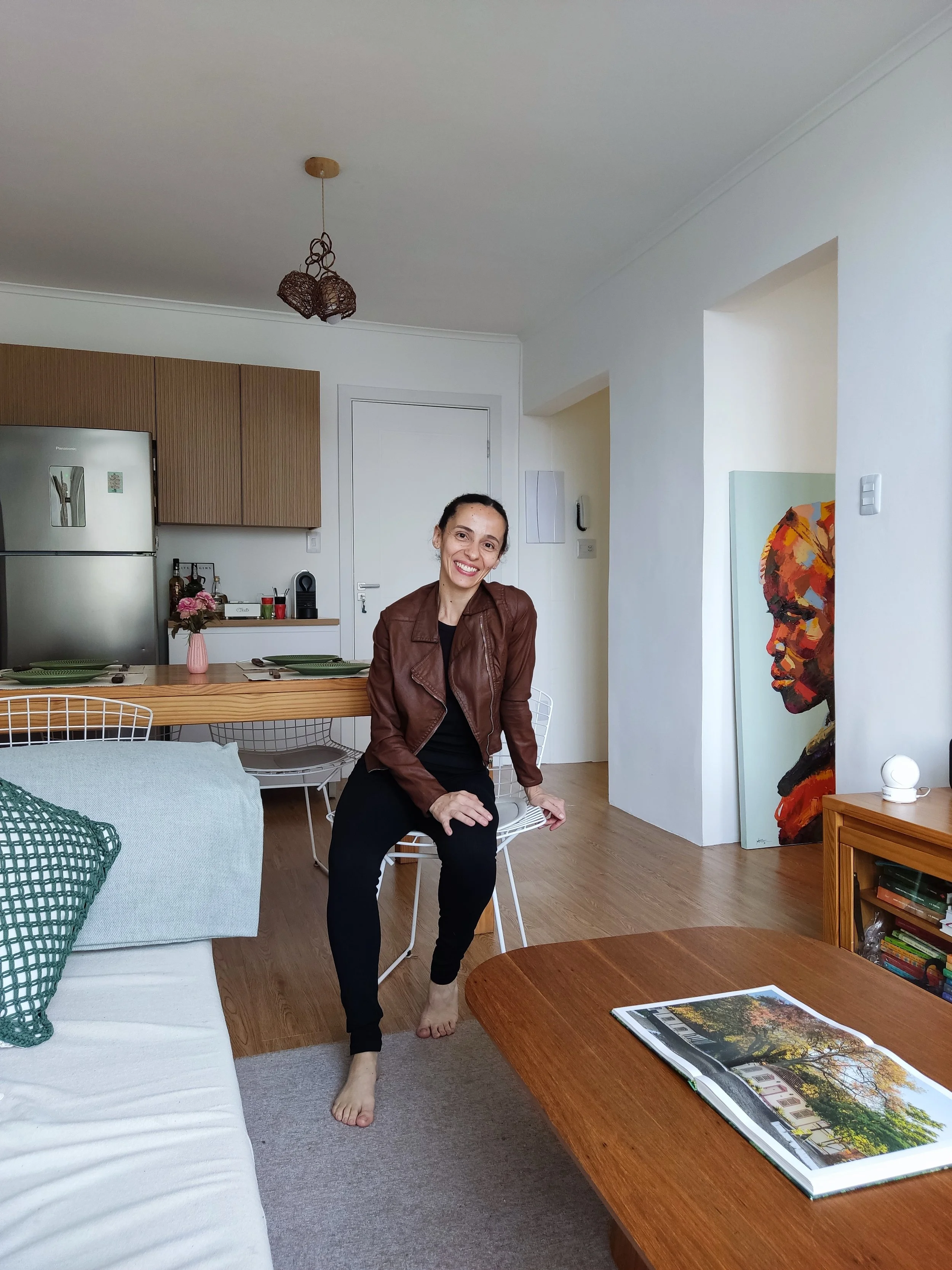 Mulher sentada em uma cadeira na sala de estar, sorrindo para a câmera, com uma parede branca ao fundo, arte moderna apoiada na parede ao lado, e uma mesa de madeira com um livro aberto com fotos de árvores e natureza.