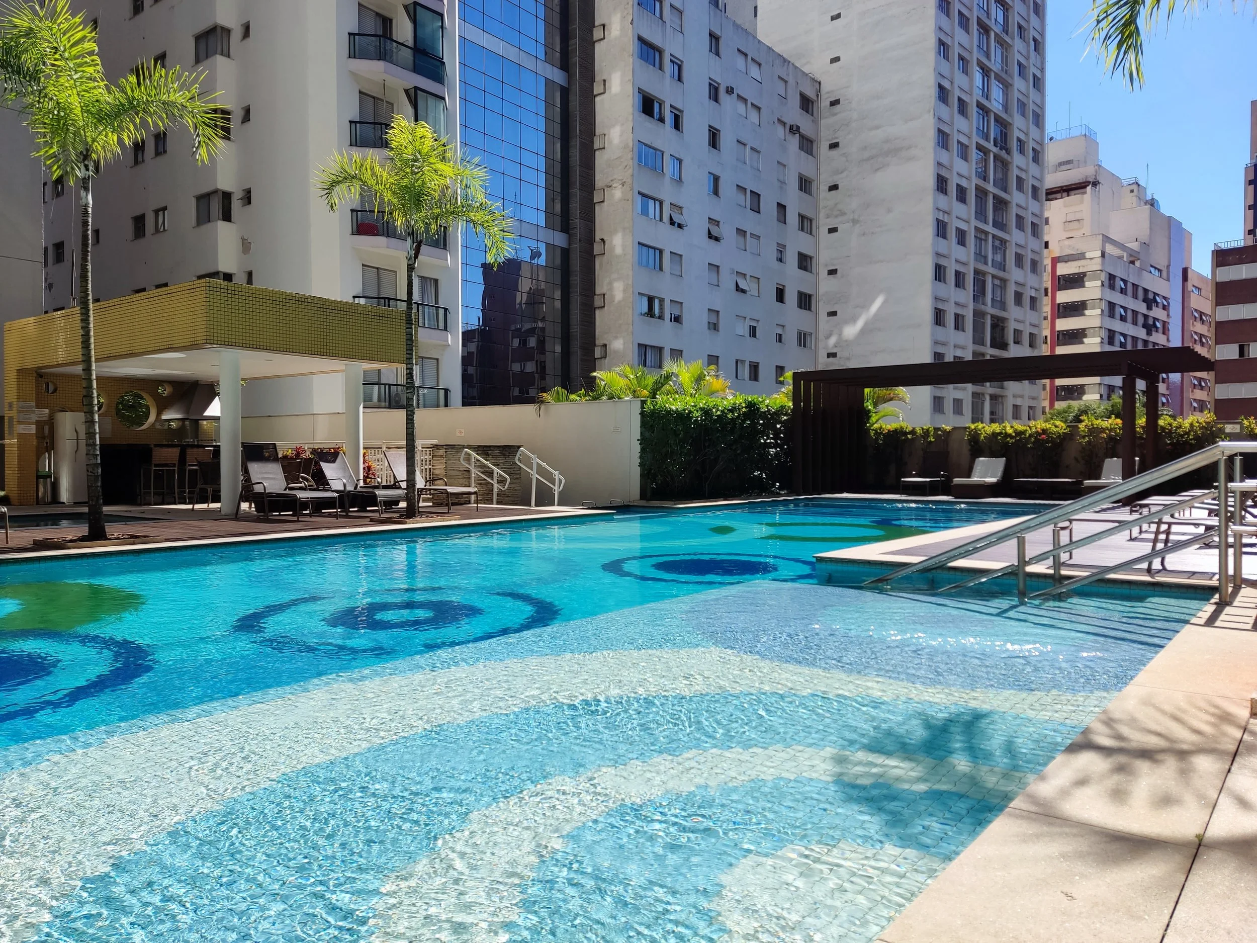 Piscina em um condomínio de prédios altos com espreguiçadeiras e área de sombra ao redor, cercada por plantas e árvores, com prédios residenciais ao fundo.