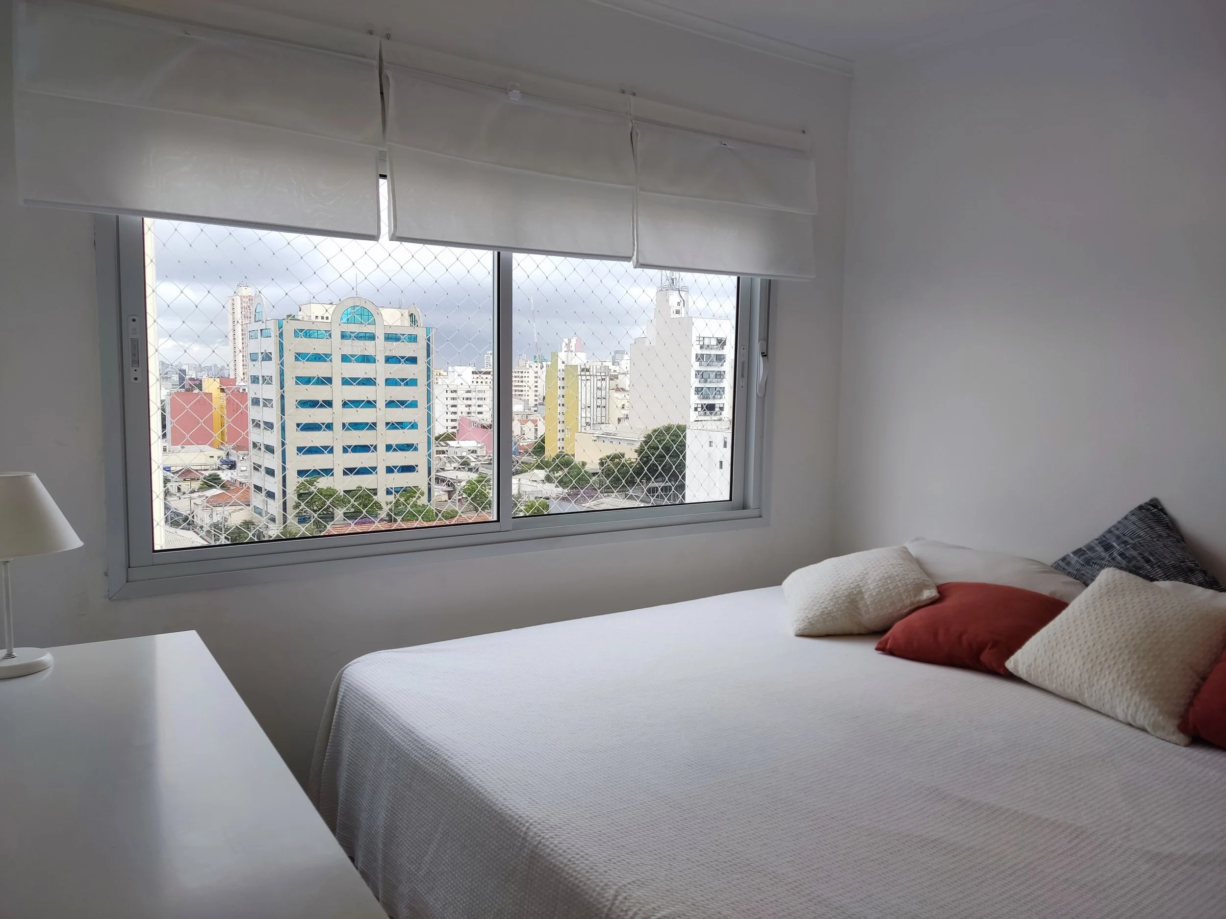 Quarto com cama de roupa branca, várias almofadas coloridas, janela com grade de proteção, vista de edifícios altos em uma cidade.