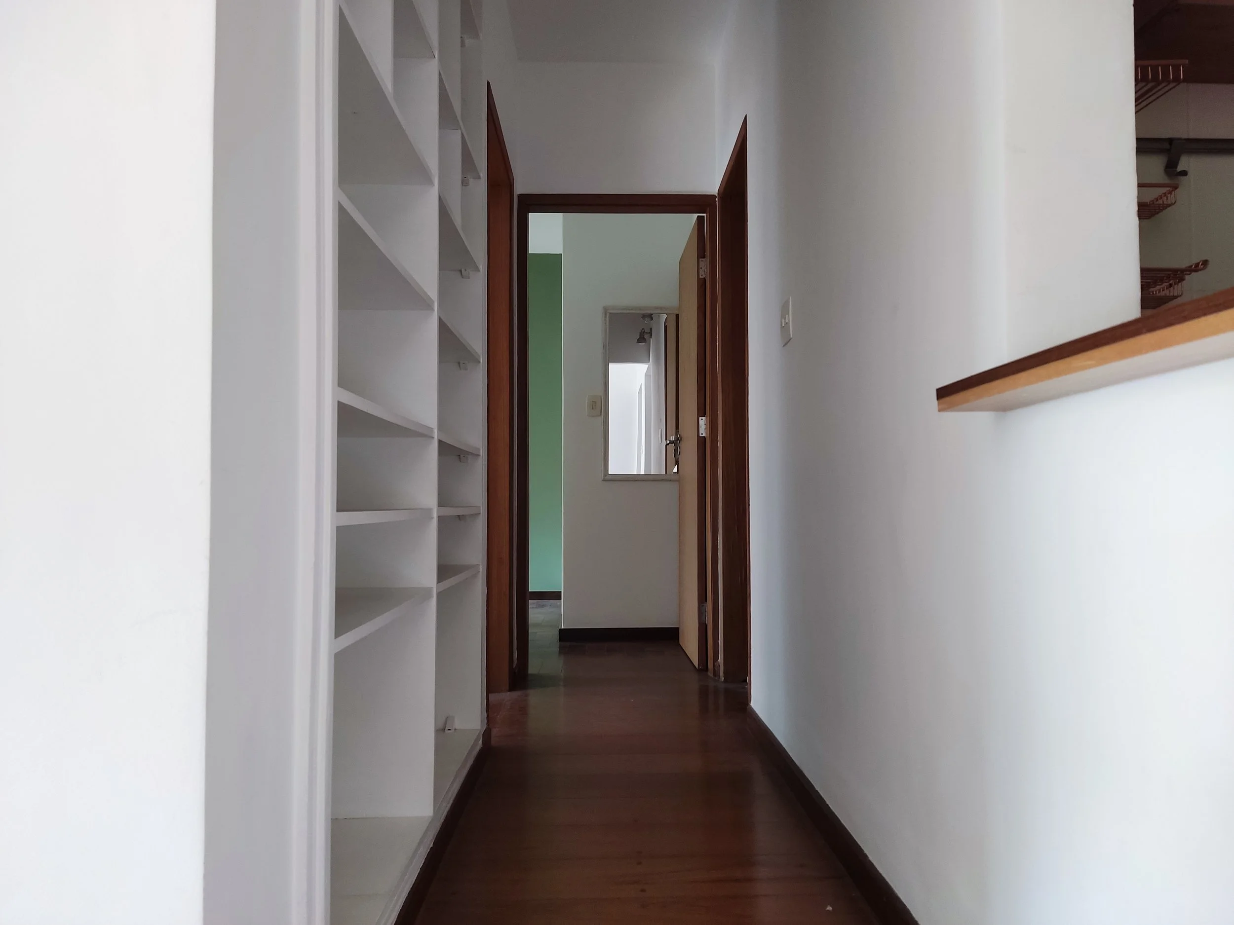Corredor com piso de madeira, paredes brancas, prateleiras brancas à esquerda, uma janela e uma porta de madeira no fundo, ao lado de uma parede lateral com um ferro para pendurar roupas.